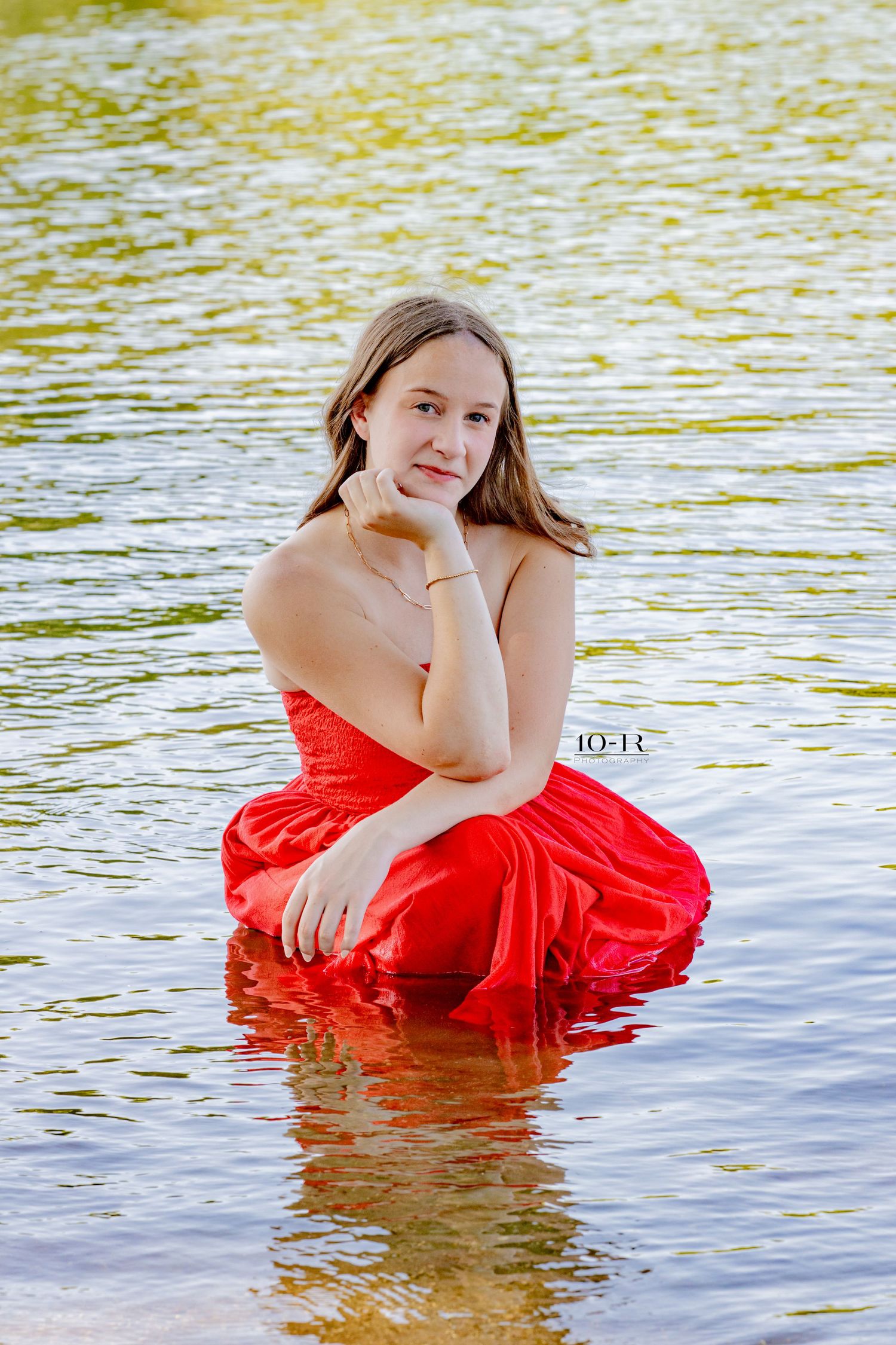 Young woman in red dress sitting in shallow water, posing gracefully with hand on chin, reflecting in calm lake.