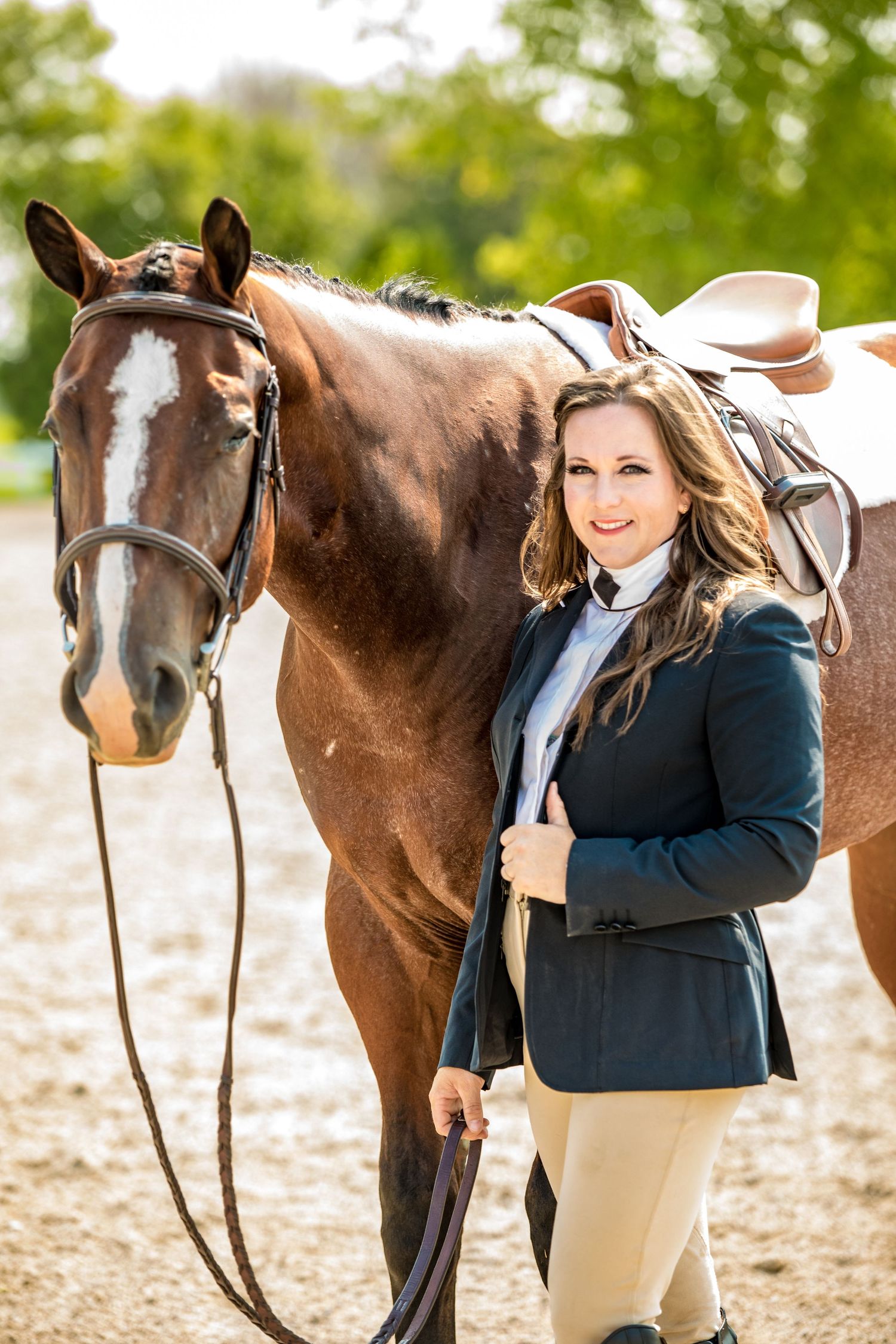 Professional equestrian in navy blazer and tan breeches poses with a brown bay horse in outdoor setting.
