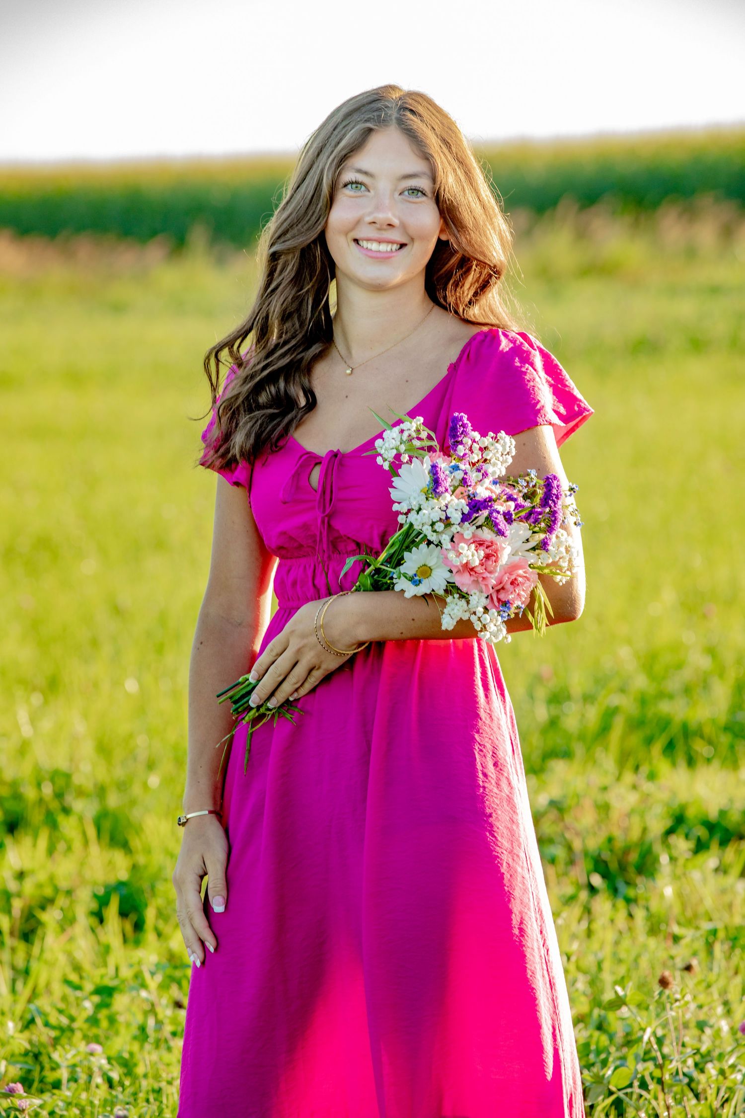 Person in vibrant pink dress holding flowers poses in sunny field during outdoor photo session.