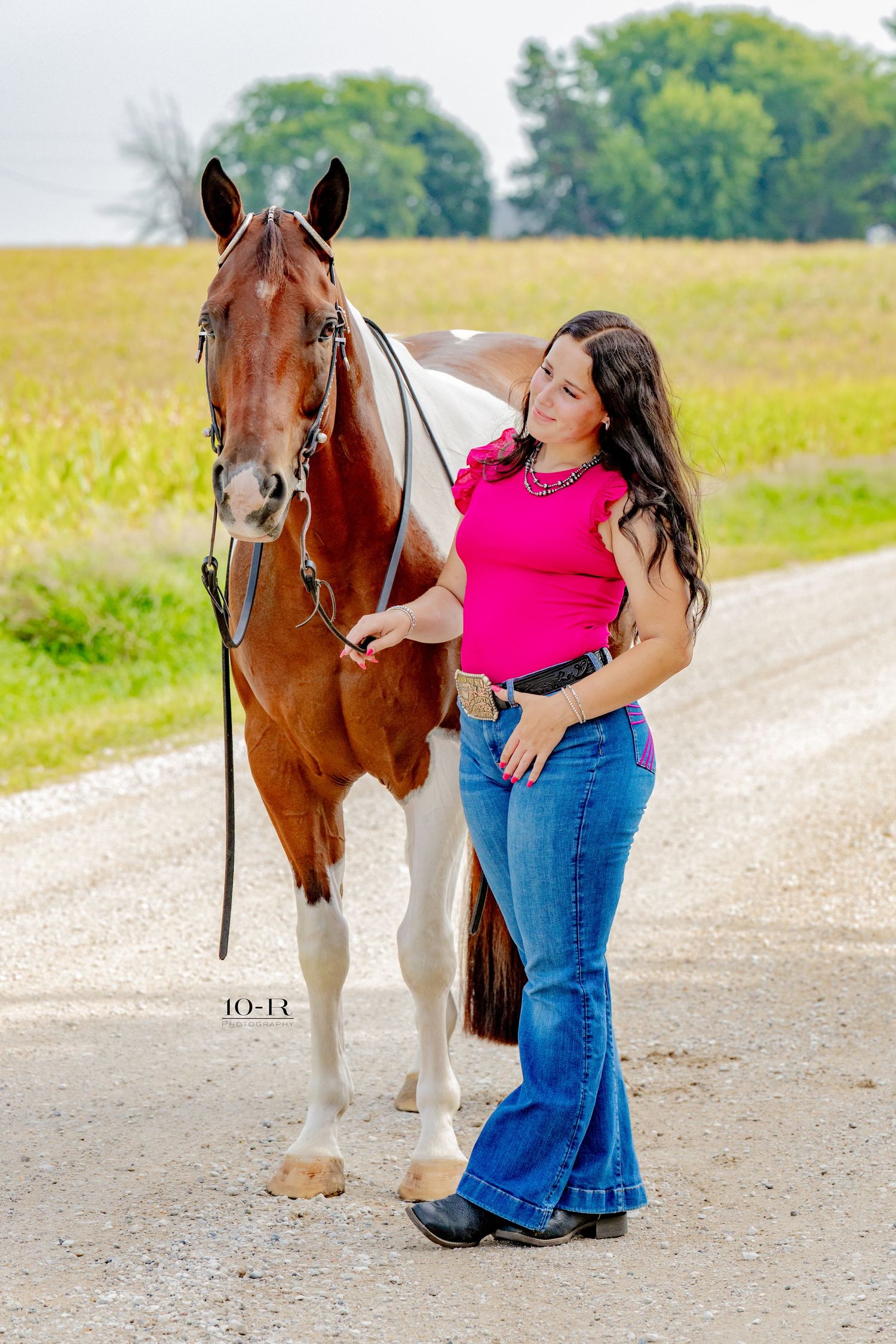 Young woman in pink top and jeans poses with a brown and white paint horse on a gravel road near farm fields.