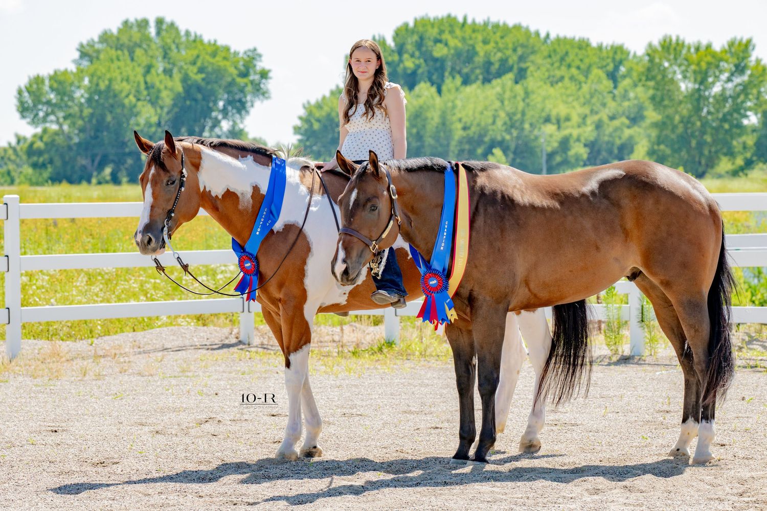 Young woman riding a bay pinto horse while leading a bay horse, both adorned with blue ribbons at an equestrian show.