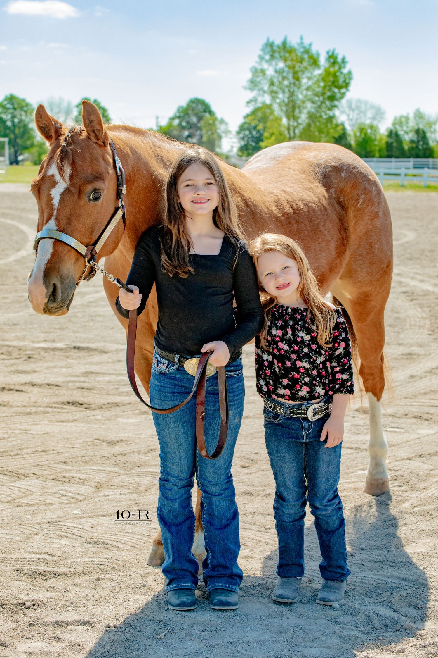 Two children standing with a beautiful chestnut horse in an outdoor riding arena on a sunny day.