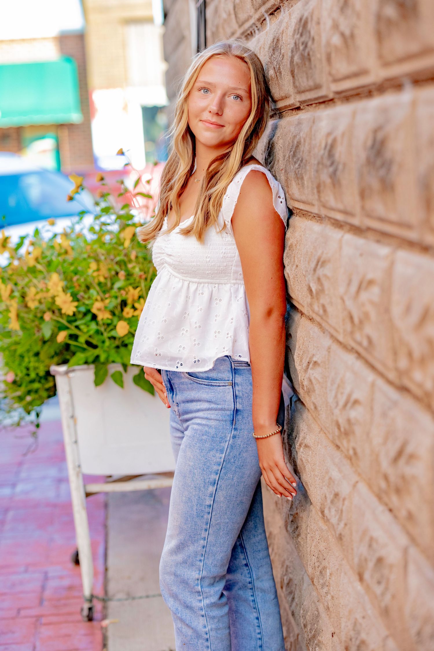 A woman wearing a frilly white summer top and light blue jeans poses casually against a stone wall near colorful flowers.