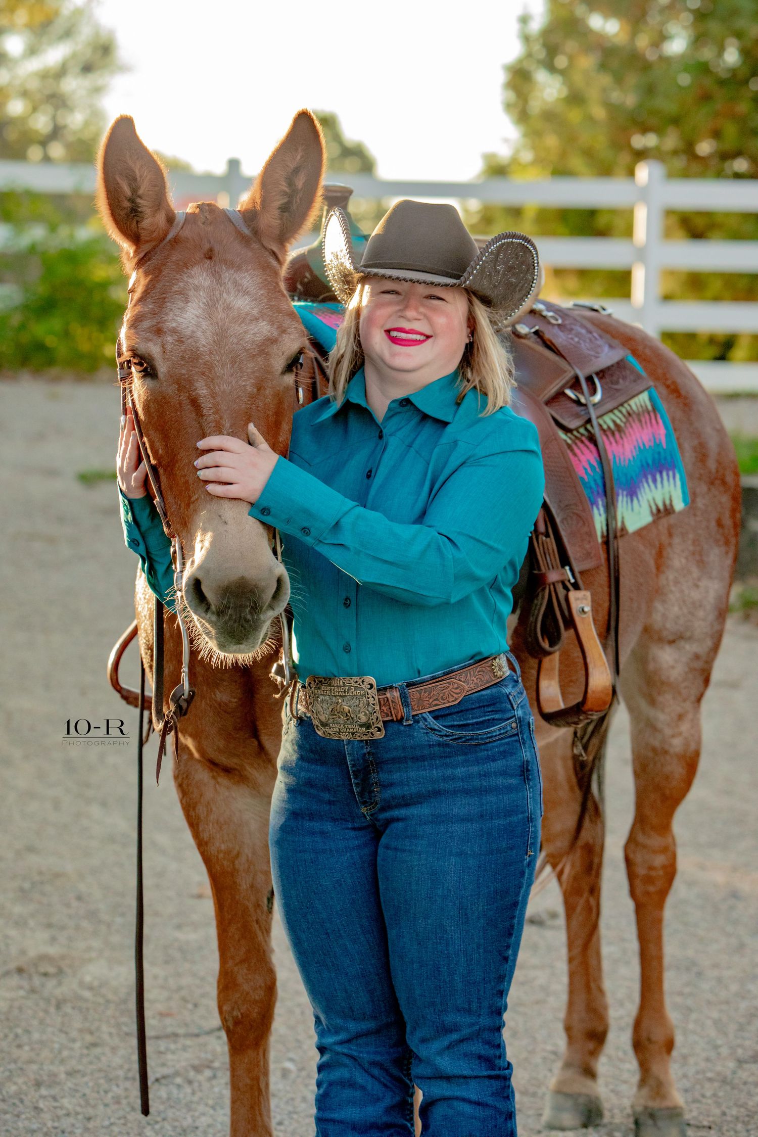 Person in western wear embracing a brown horse while smiling in an outdoor arena.