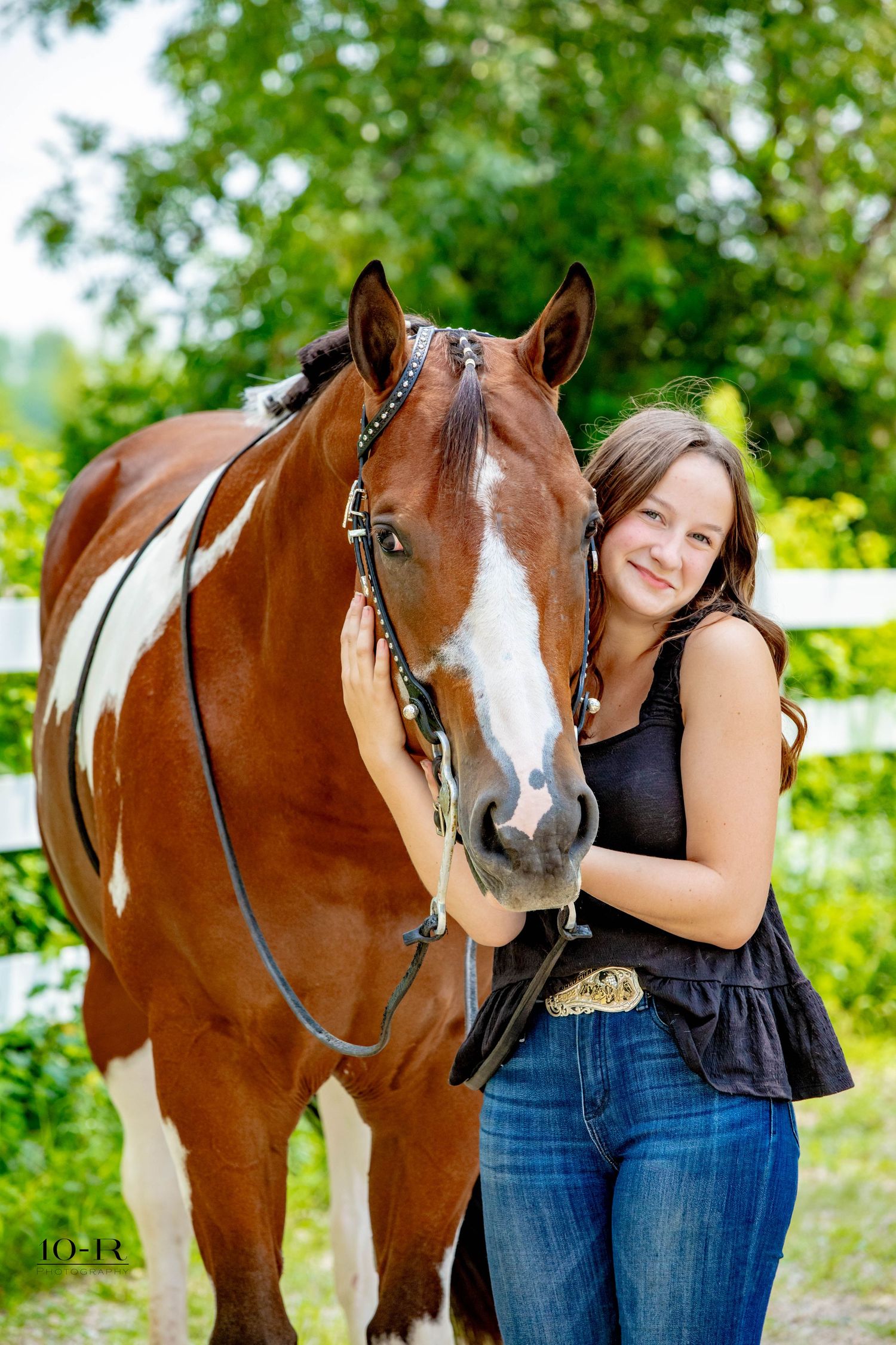 A paint horse with distinctive brown and white markings poses in front of green trees.