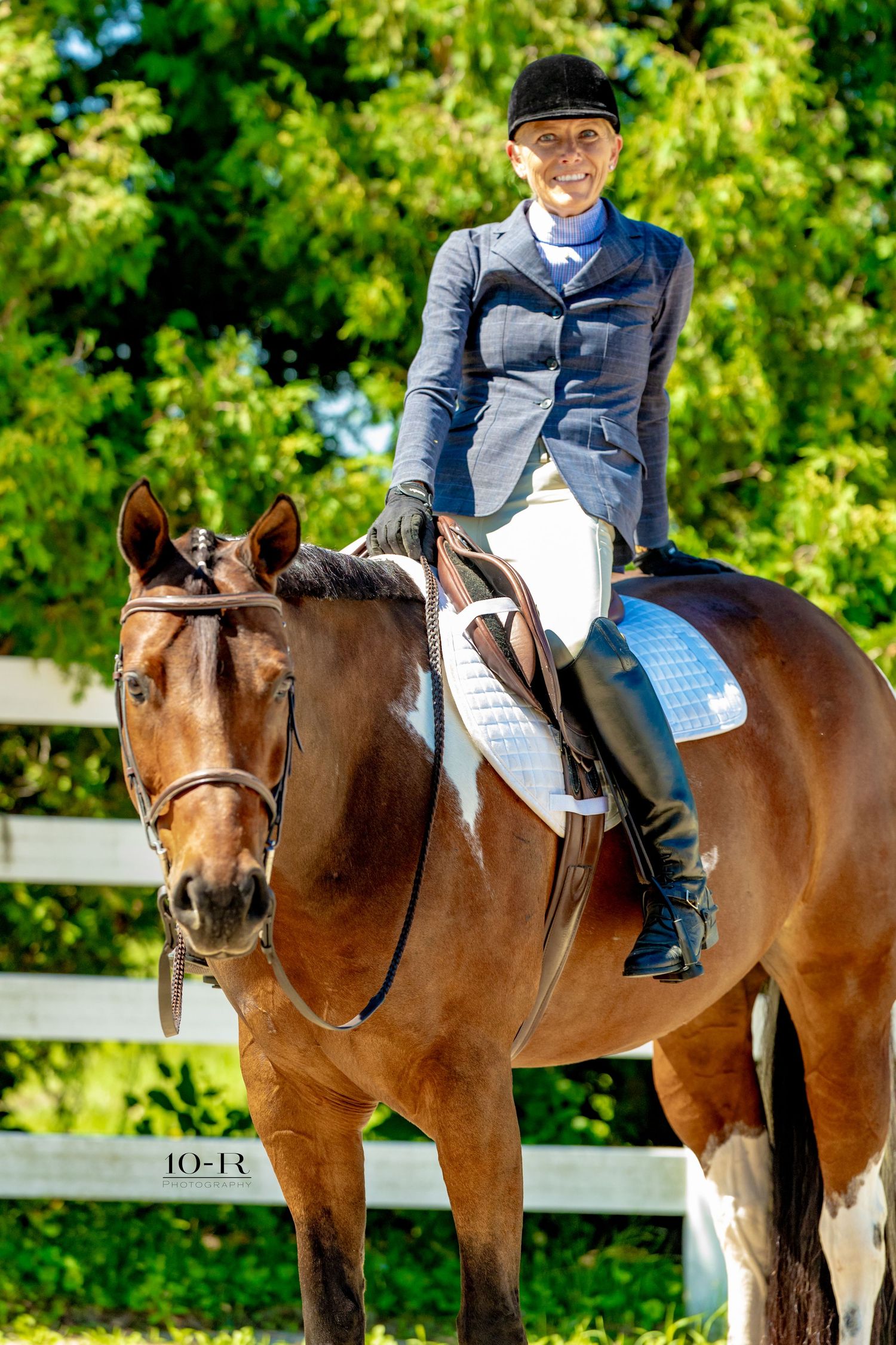 Smiling equestrian in blue jacket and white breeches riding a chestnut horse at an outdoor dressage competition.