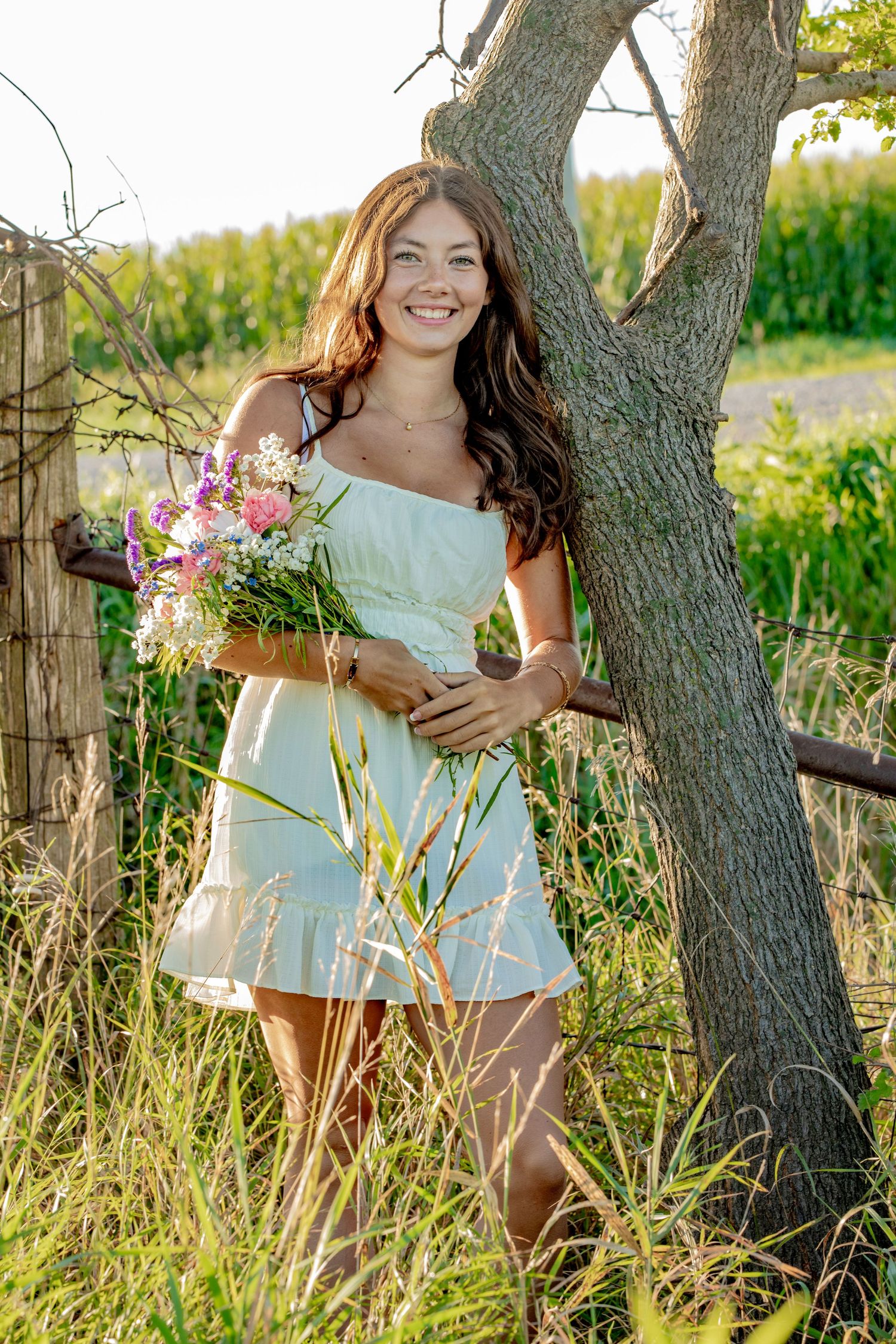 Woman in white dress poses against tree trunk in golden sunlit meadow during summer photoshoot.