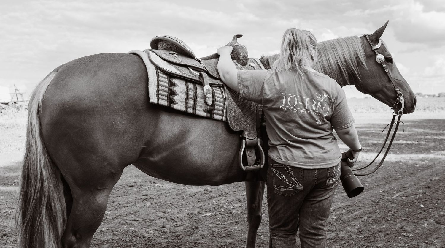 A woman in a gray t-shirt stands beside a saddled brown horse in an open field, viewed from behind.