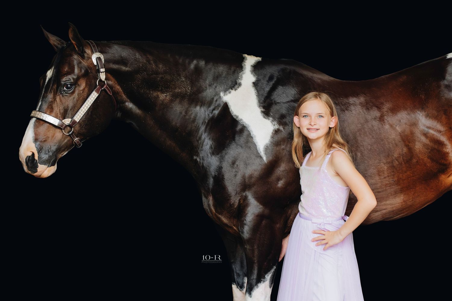 A young girl in a white dress and brown boots stands confidently next to a dark brown horse outdoors.
