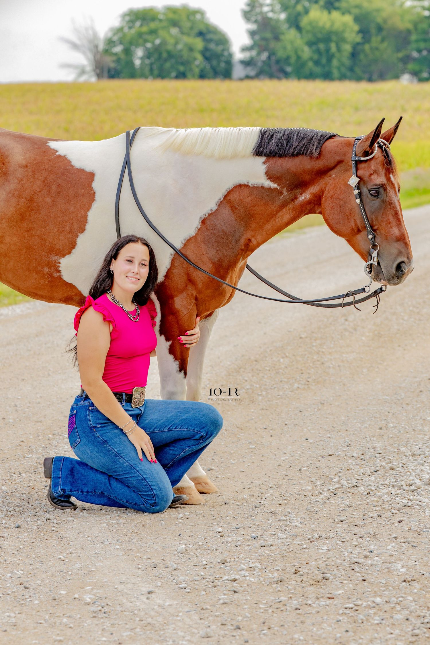 Woman in pink top and jeans kneels beside a brown and white paint horse on a gravel path.