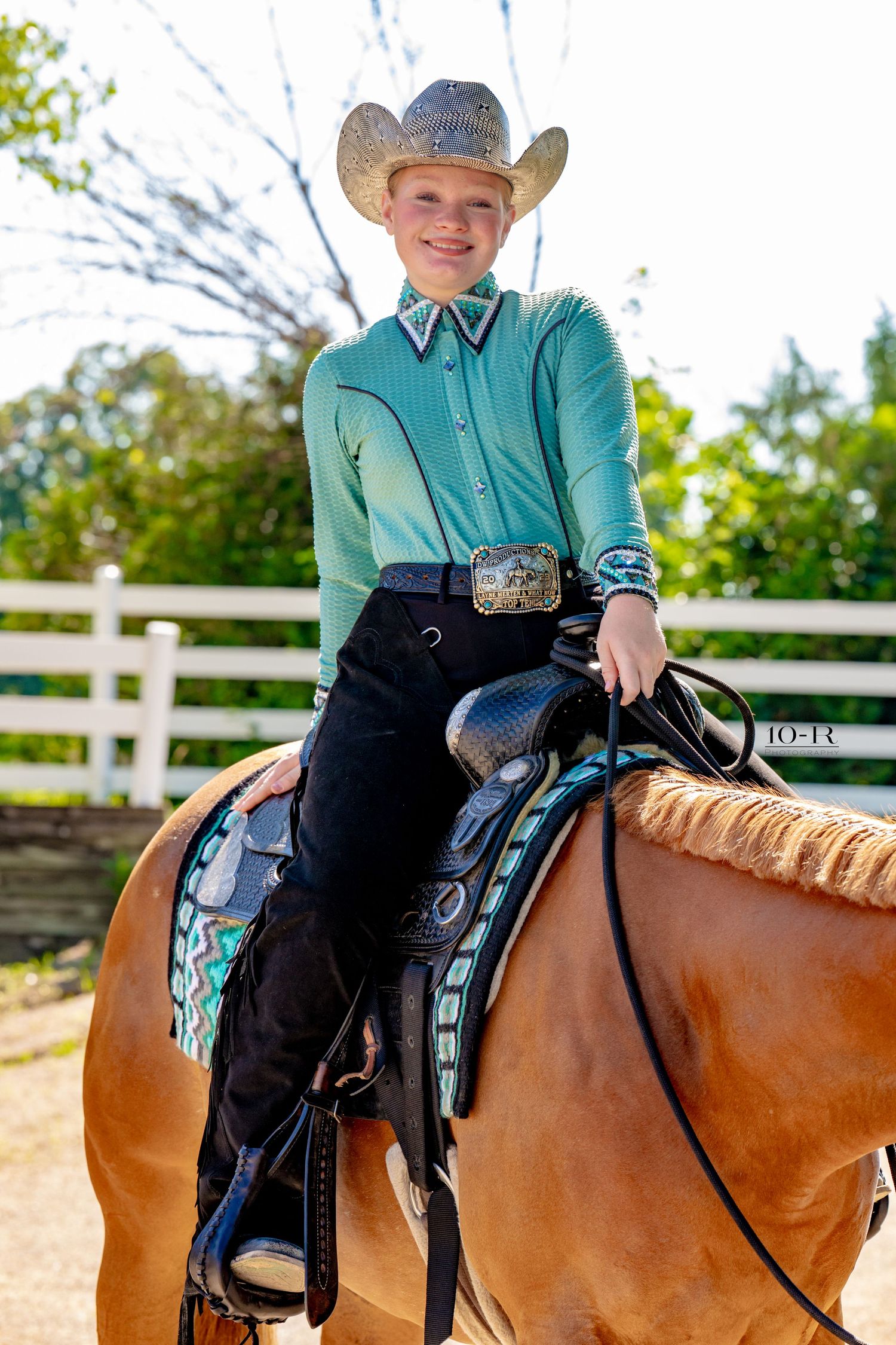 Young rider in turquoise western shirt and cowboy hat smiles atop a palomino horse with decorative saddle pad.