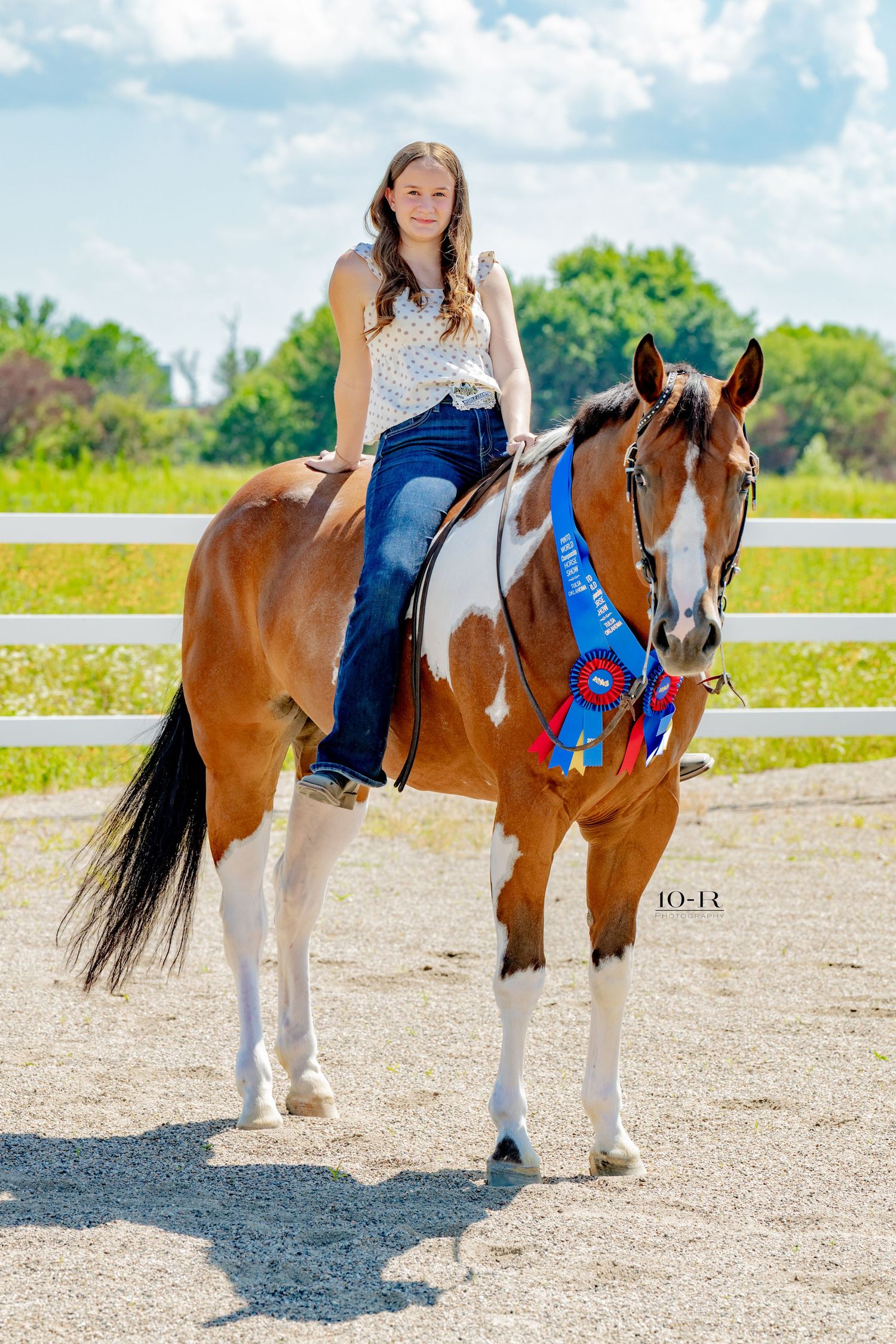 Young woman riding a chestnut paint horse with blue ribbon award at an outdoor equestrian show.