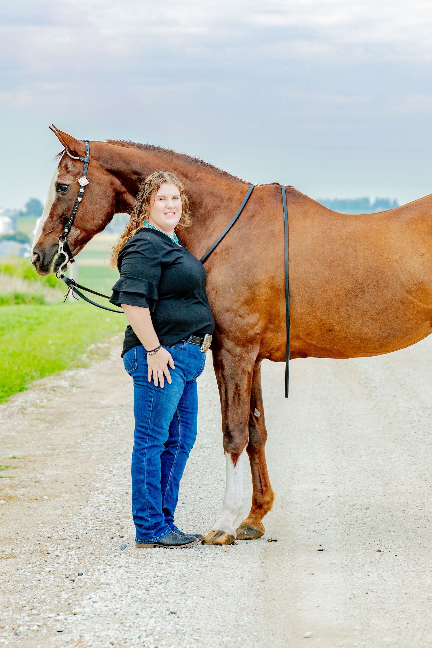 A smiling woman in a black top and jeans stands beside a chestnut horse on a gravel path with green fields behind.