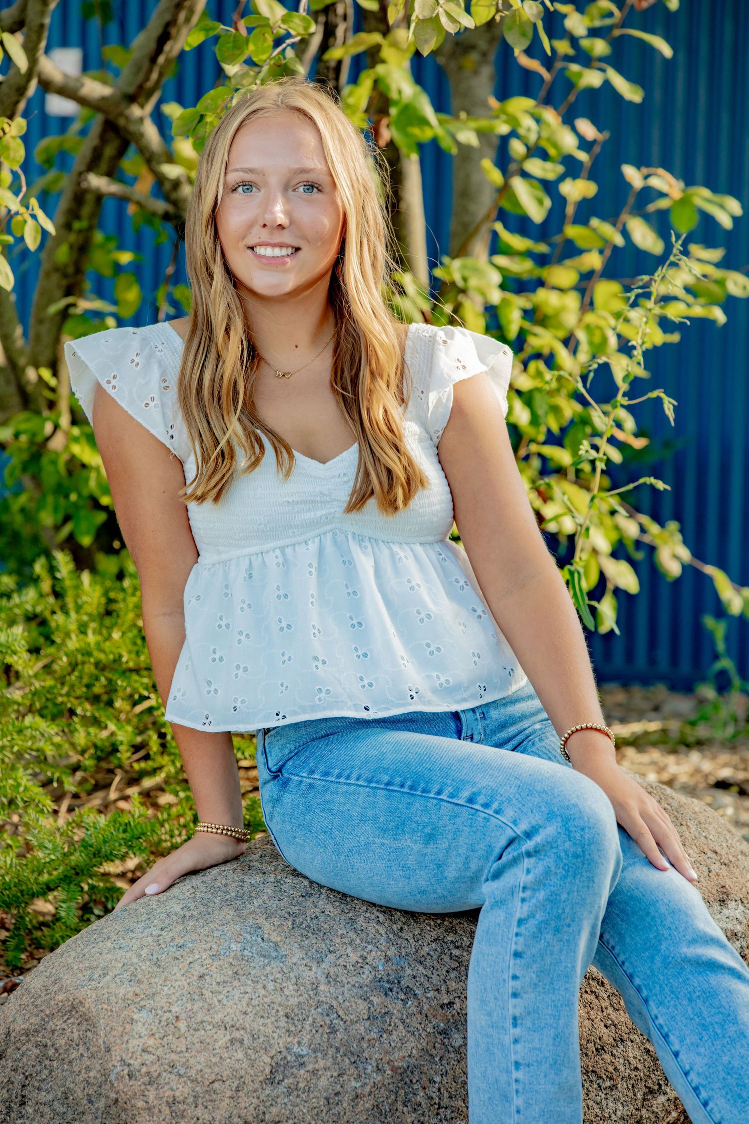 A casual summer portrait on a rock wearing a white eyelet top and blue jeans against green foliage.