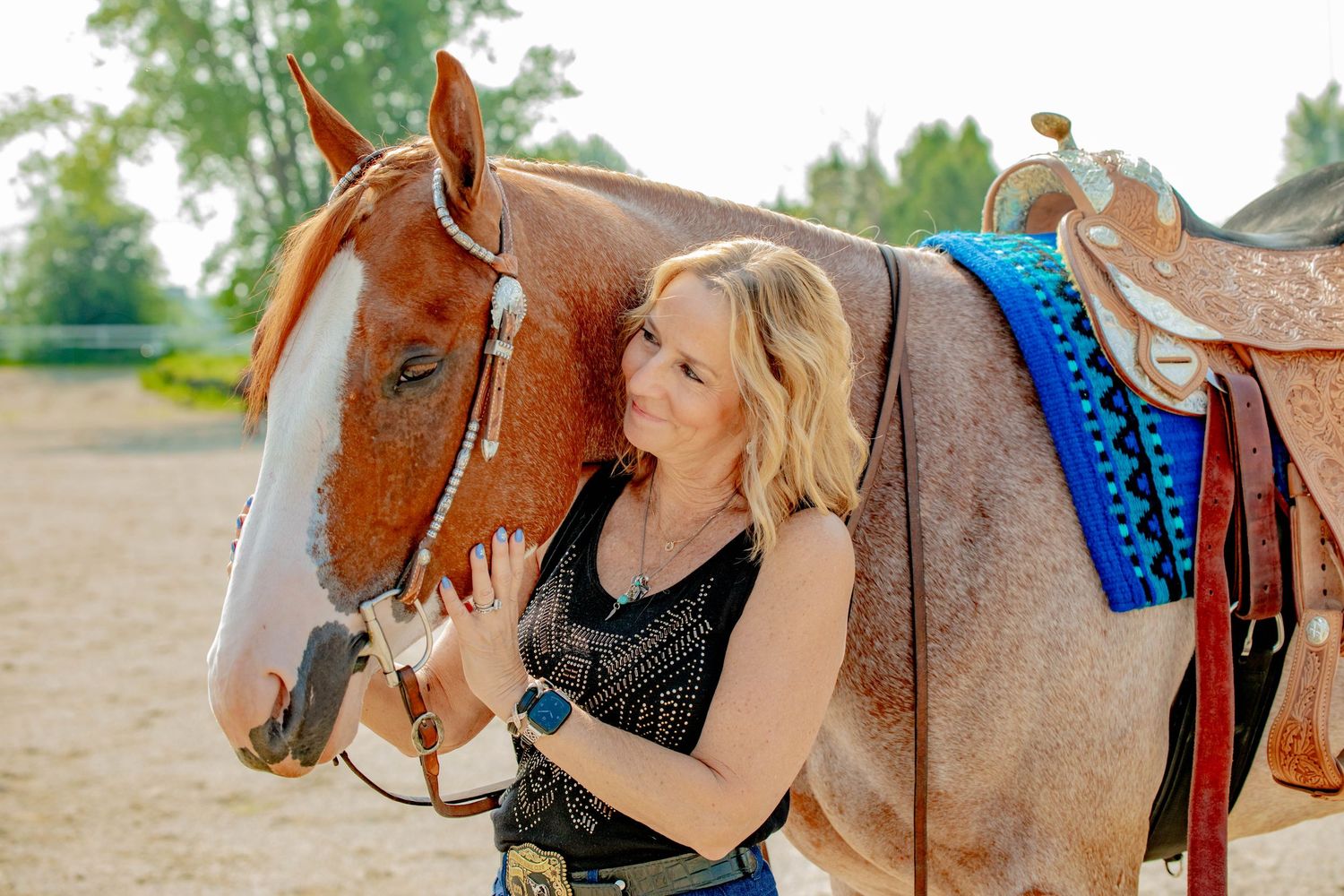 A chestnut horse with white markings wearing a western saddle and blue pad stands calmly in a sunny outdoor setting.