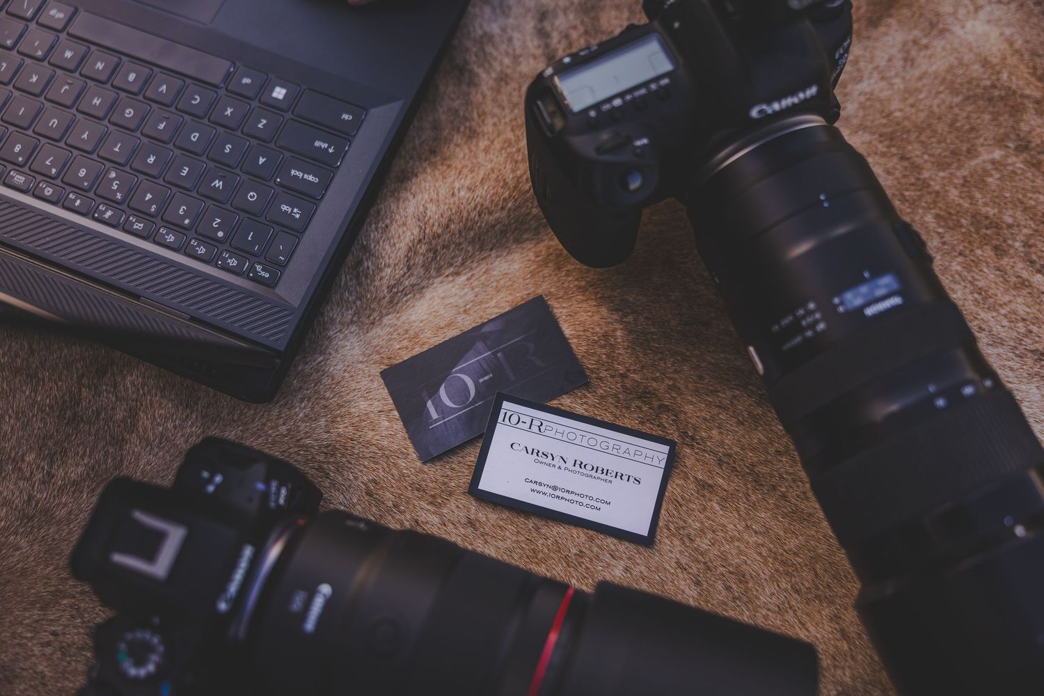 Two Canon DSLR cameras with lenses beside a laptop and business card on a textured surface.