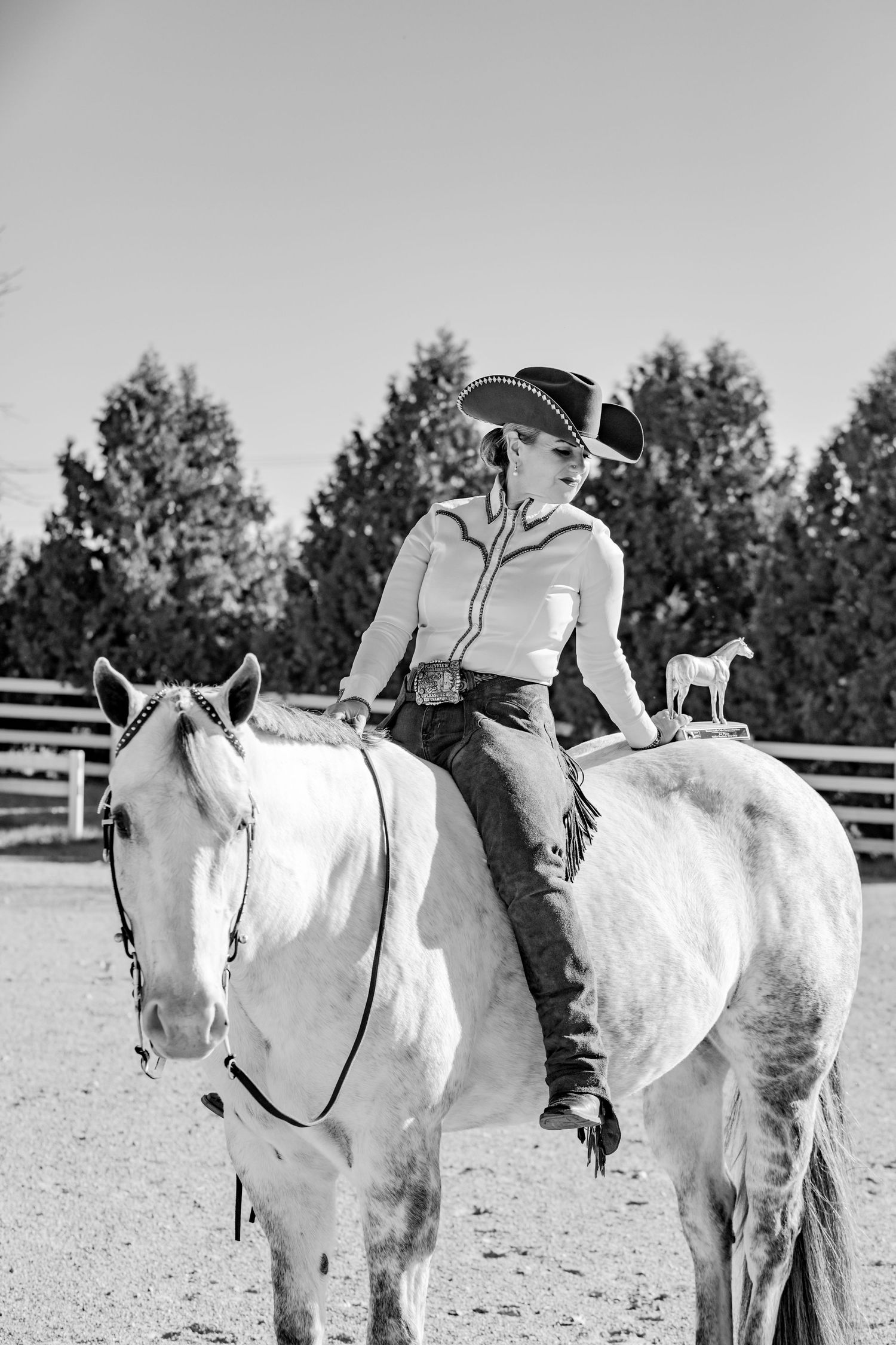 Black and white photo of a rider in western attire sitting on a white horse in a fenced arena with trees in background.