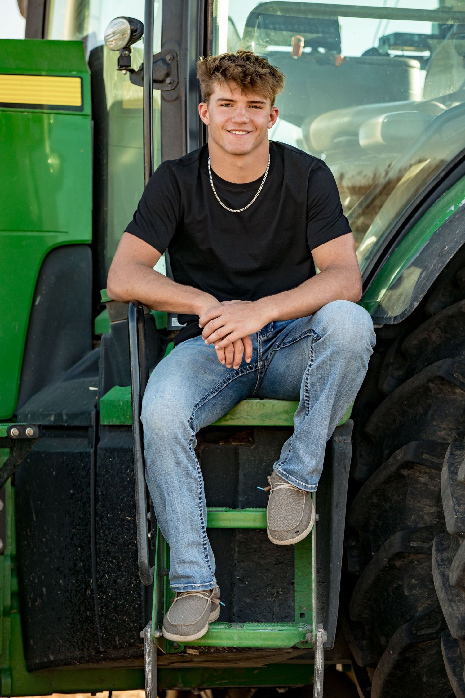 Person sits casually on steps of green tractor wearing black t-shirt and jeans.
