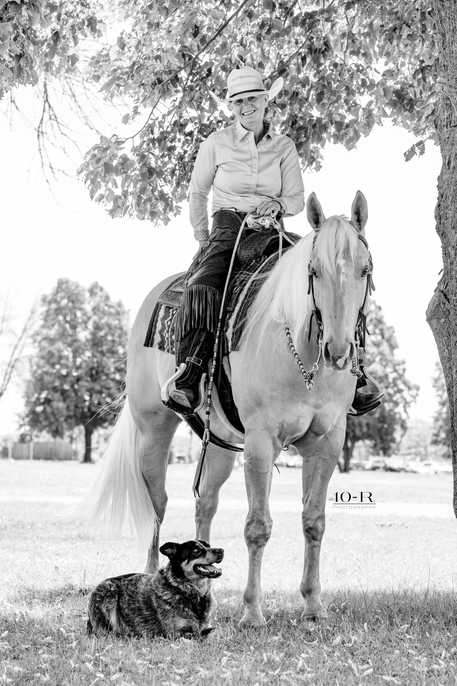 Black and white photo of a smiling woman in cowboy hat riding a white horse with a dog resting nearby under a tree.