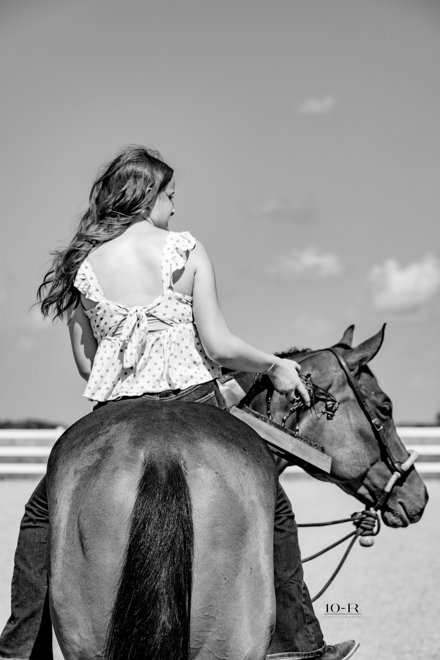 Black and white photo of a woman in a backless top riding a horse under a cloudy sky.
