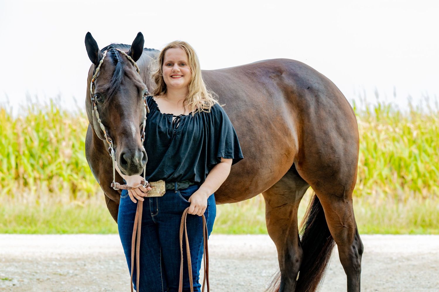 Woman in black outfit standing beside a dark bay horse outdoors near green fields.