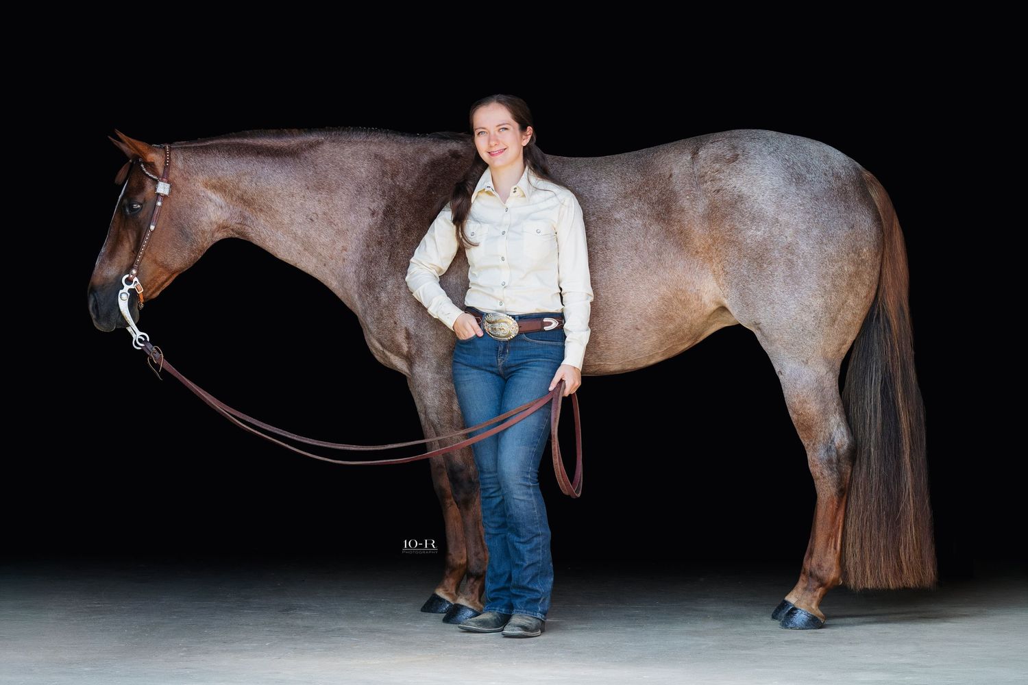 Woman in western attire standing beside a red roan horse against a dark background.