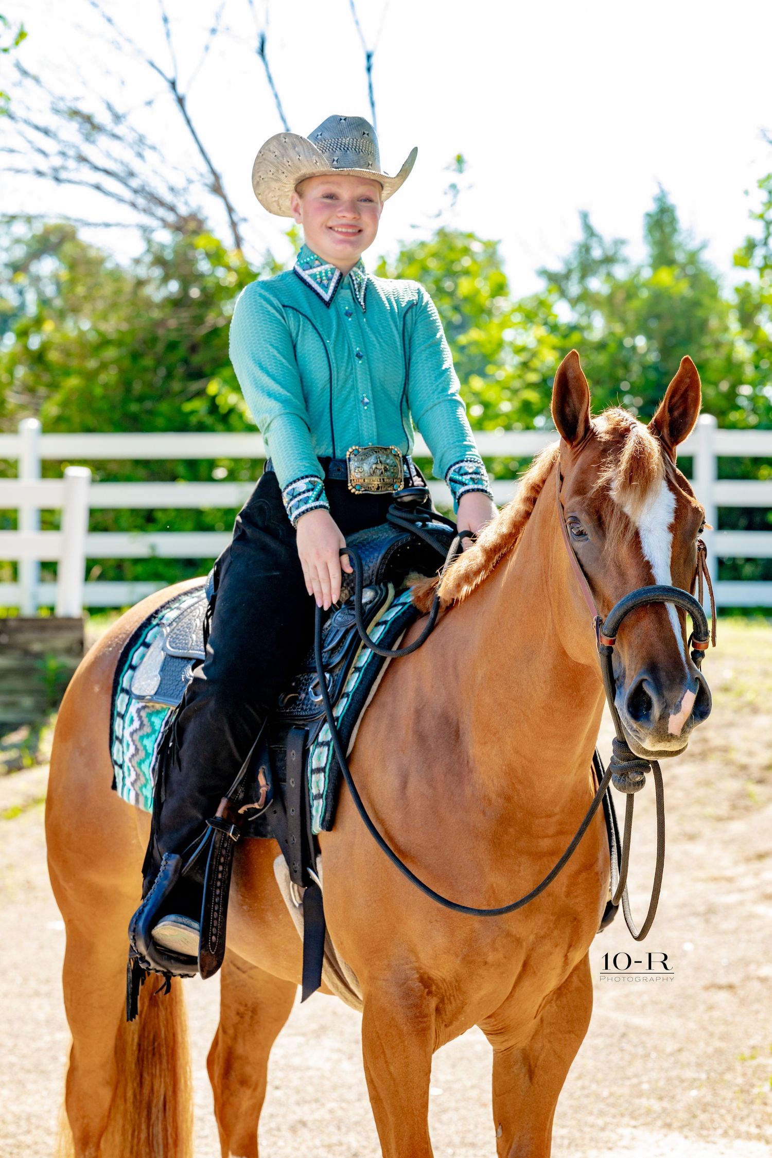 Young rider in turquoise shirt and cowboy hat sits atop a chestnut horse in a white-fenced arena.