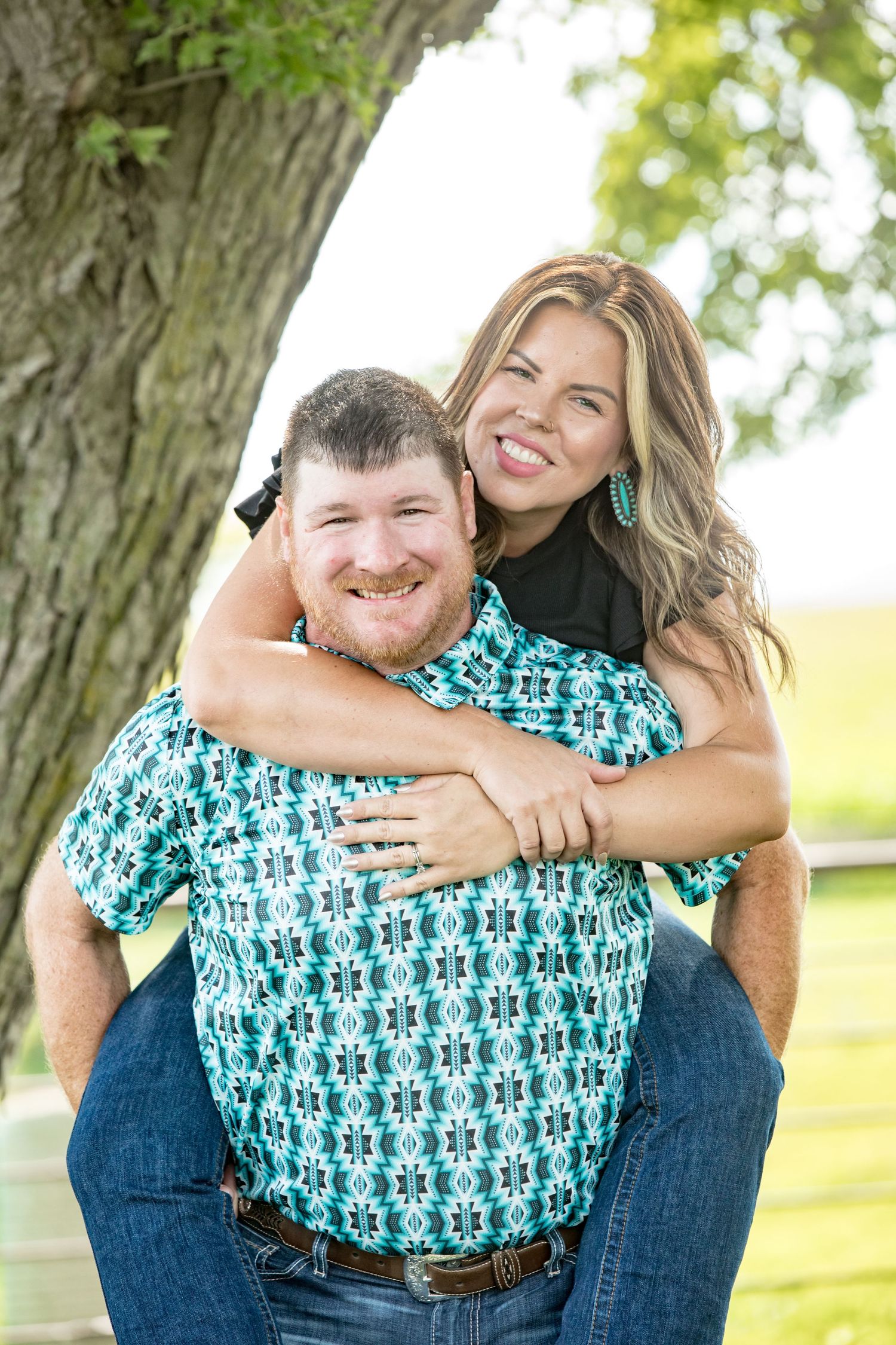 Playful couple sharing a piggyback ride moment outdoors under a large tree.