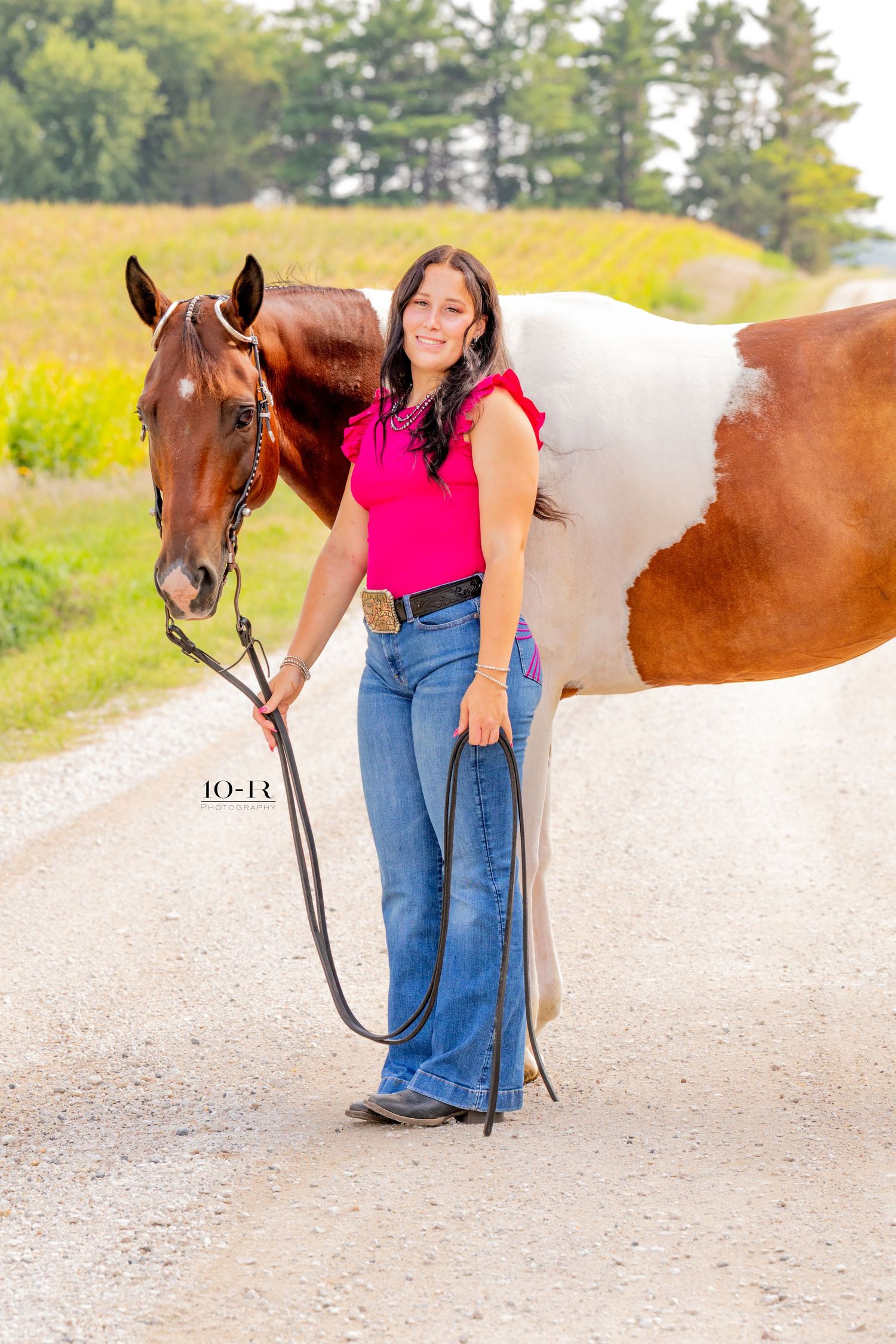 A young woman in a pink top and jeans holds the reins of a brown and white paint horse on a gravel road.