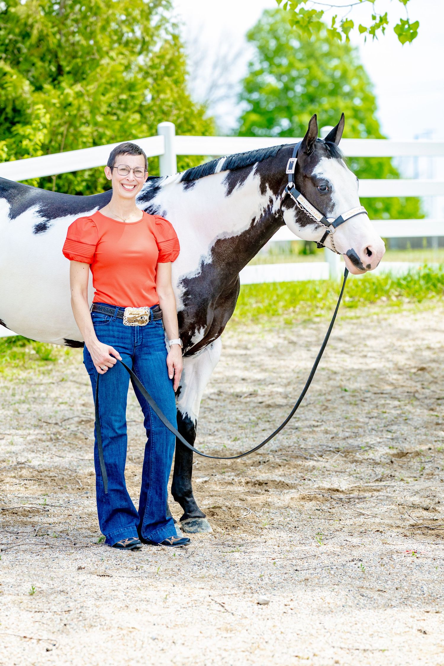 Woman in red top and jeans smiling beside a black and white paint horse at an outdoor equestrian facility.