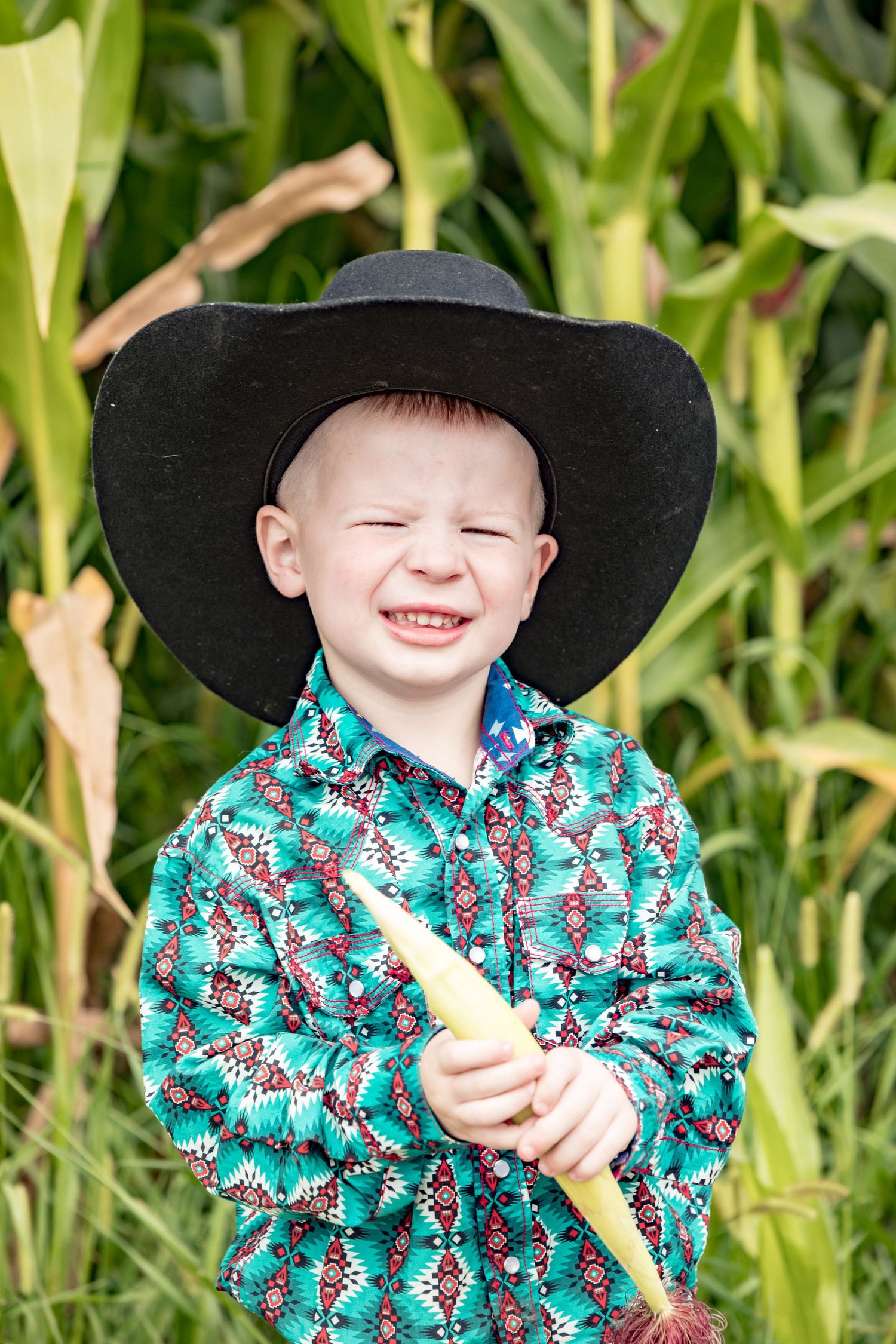 Young child in a cowboy hat and patterned shirt smiling in front of tall corn stalks.
