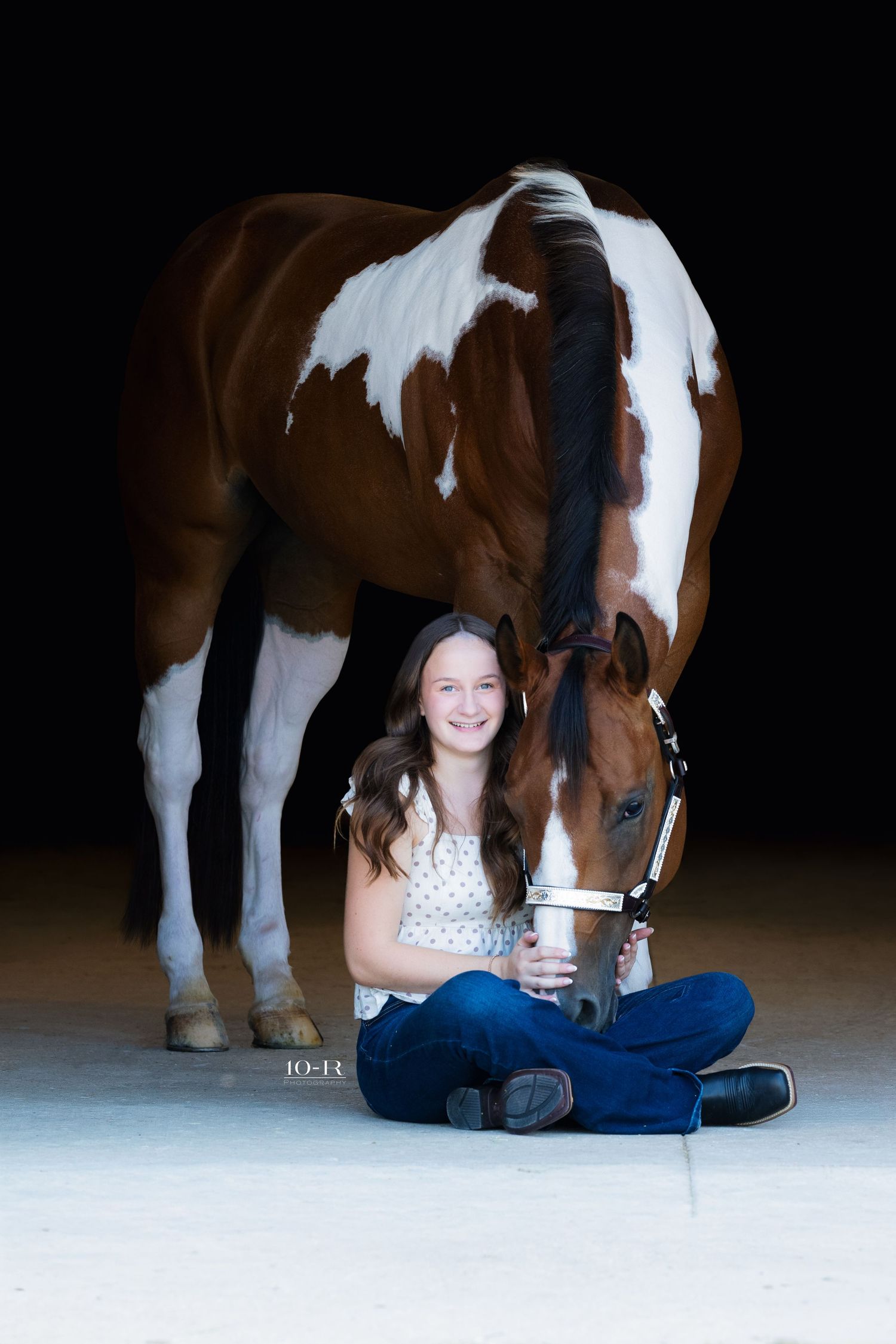 Smiling teenage girl sitting on floor beside a brown and white paint horse in dark studio.