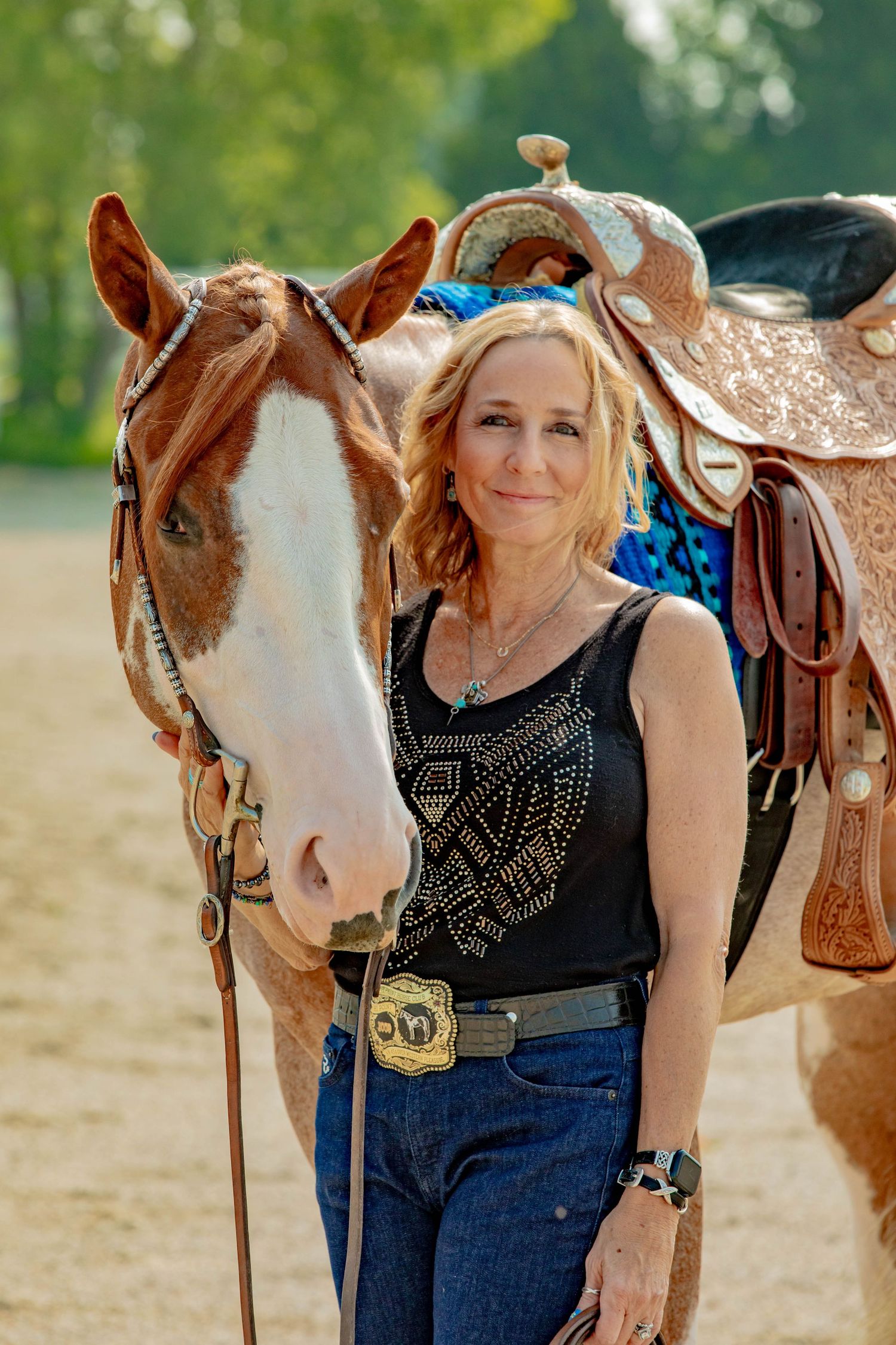 A brown and white horse stands next to a Western saddle with its rider in a black studded top and blue jeans.