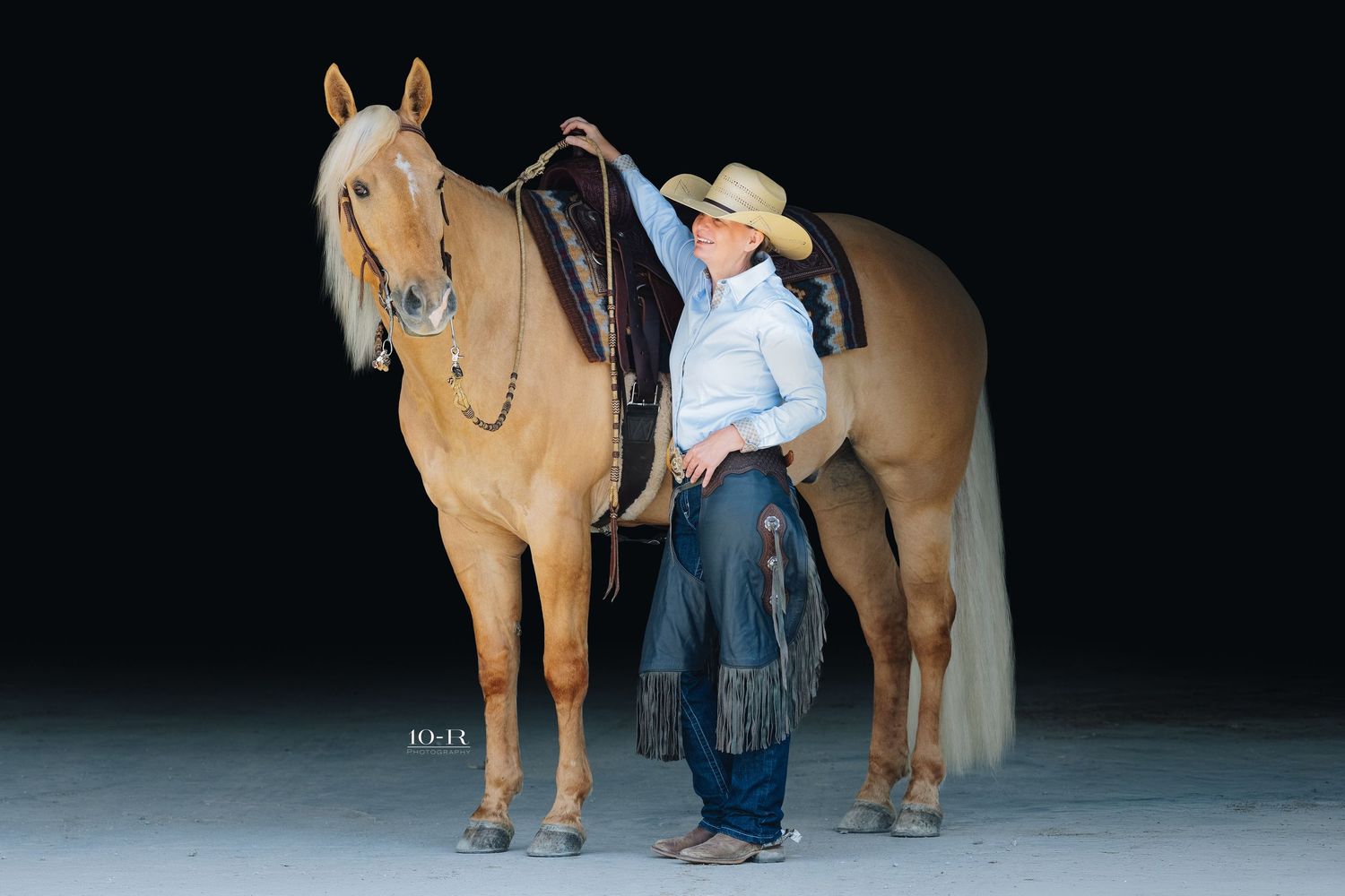 A cowgirl in a white shirt and hat stands beside a golden palomino horse with a Western saddle on a dark background.