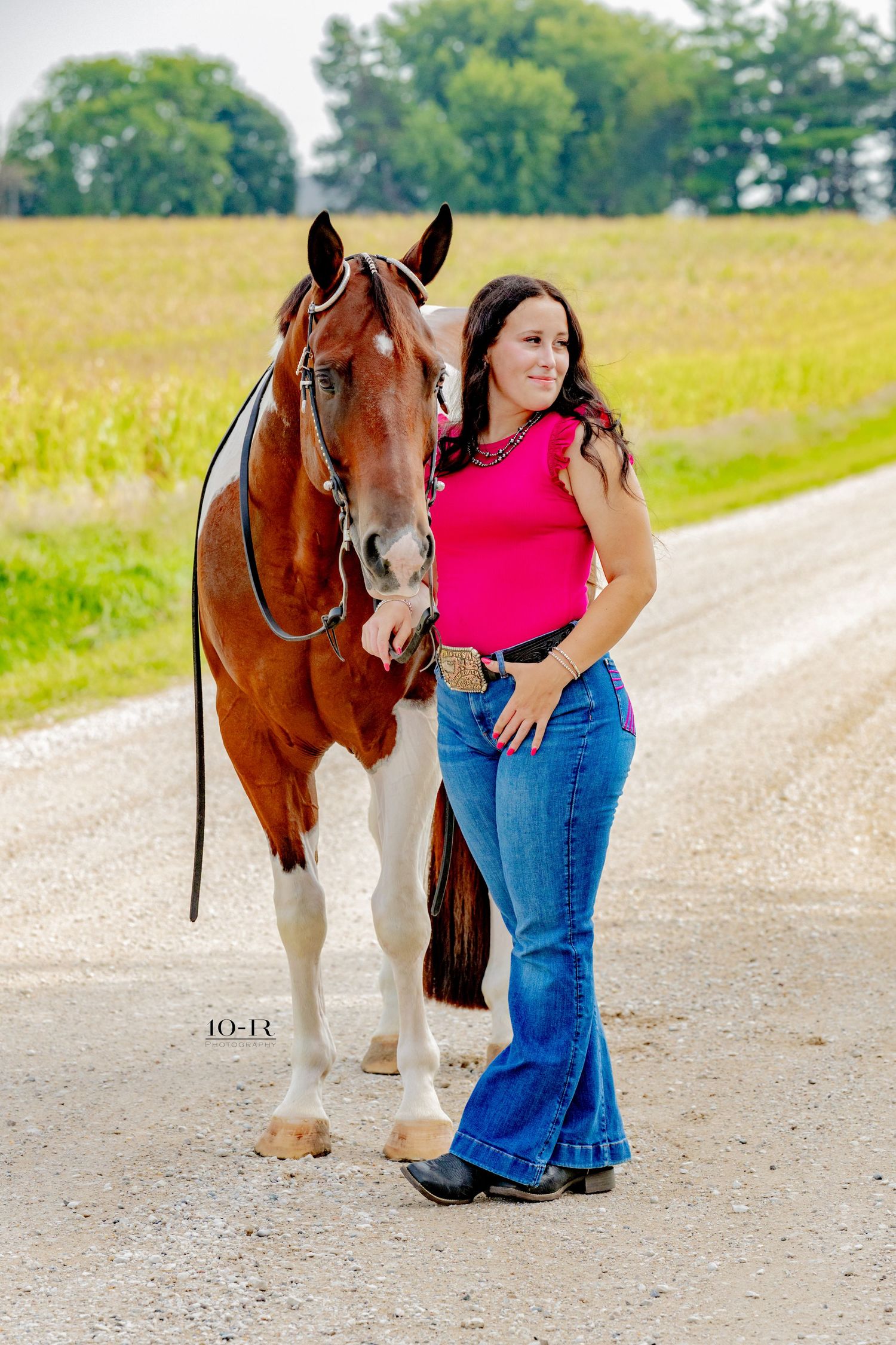 Woman in pink top and jeans poses with a chestnut paint horse on a gravel road beside green fields.