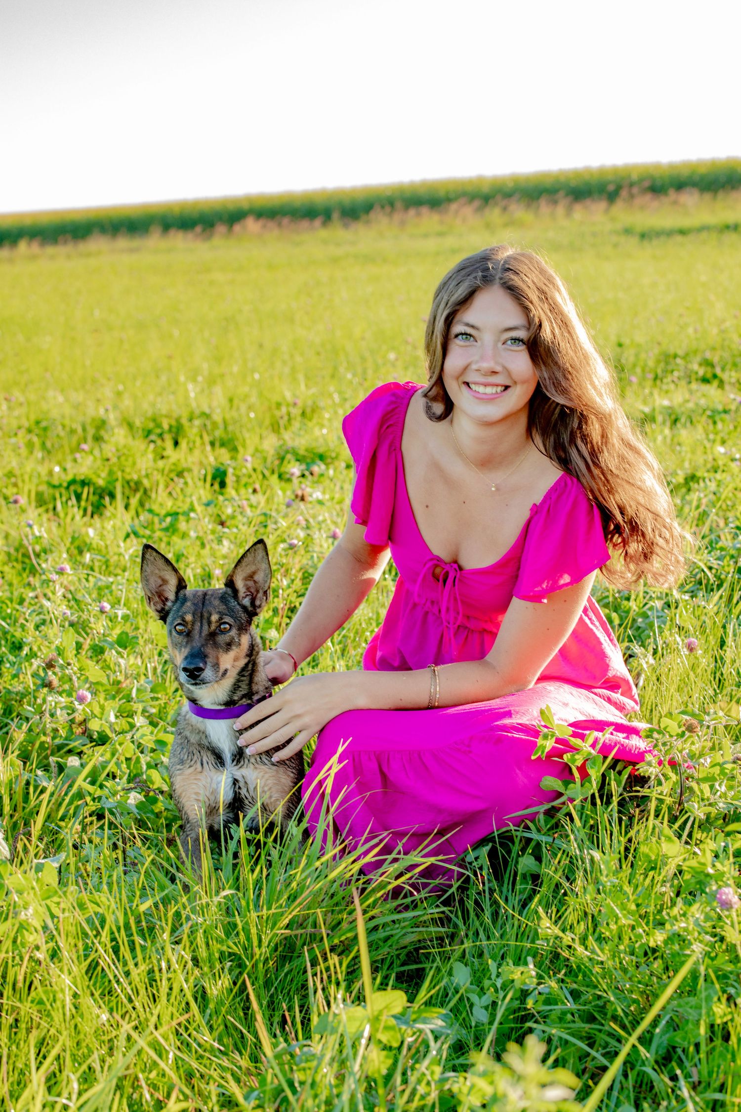 Person in bright pink dress sitting in sunny meadow with small black and white dog on green grass.