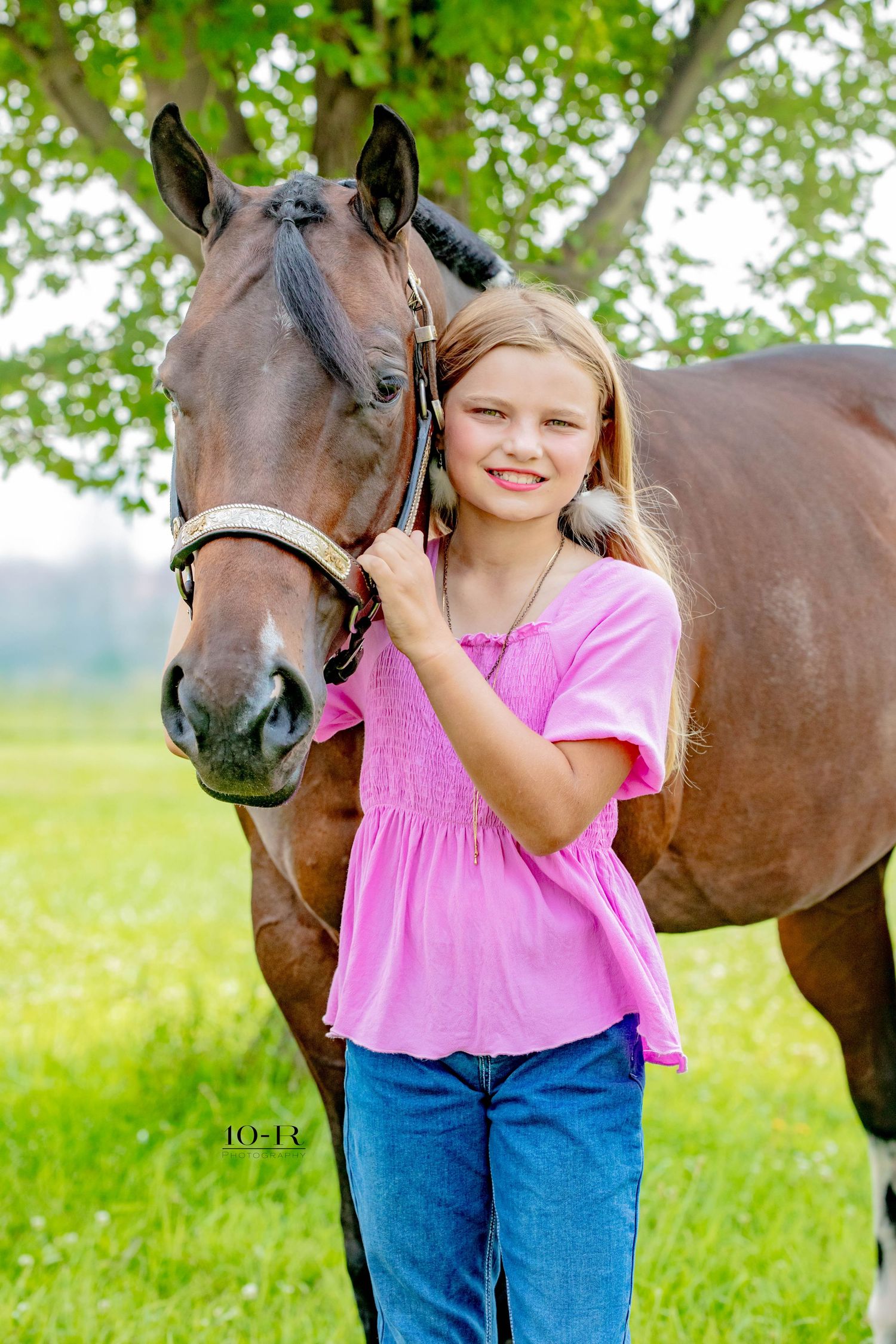 A girl in a pink shirt poses with her bay horse on a sunny summer day in a grassy field.