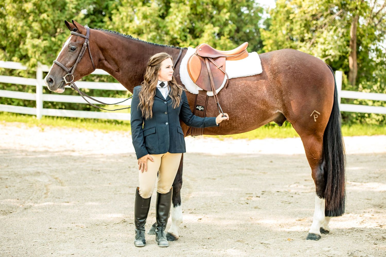 English rider in full show attire stands beside bay horse near white fence.