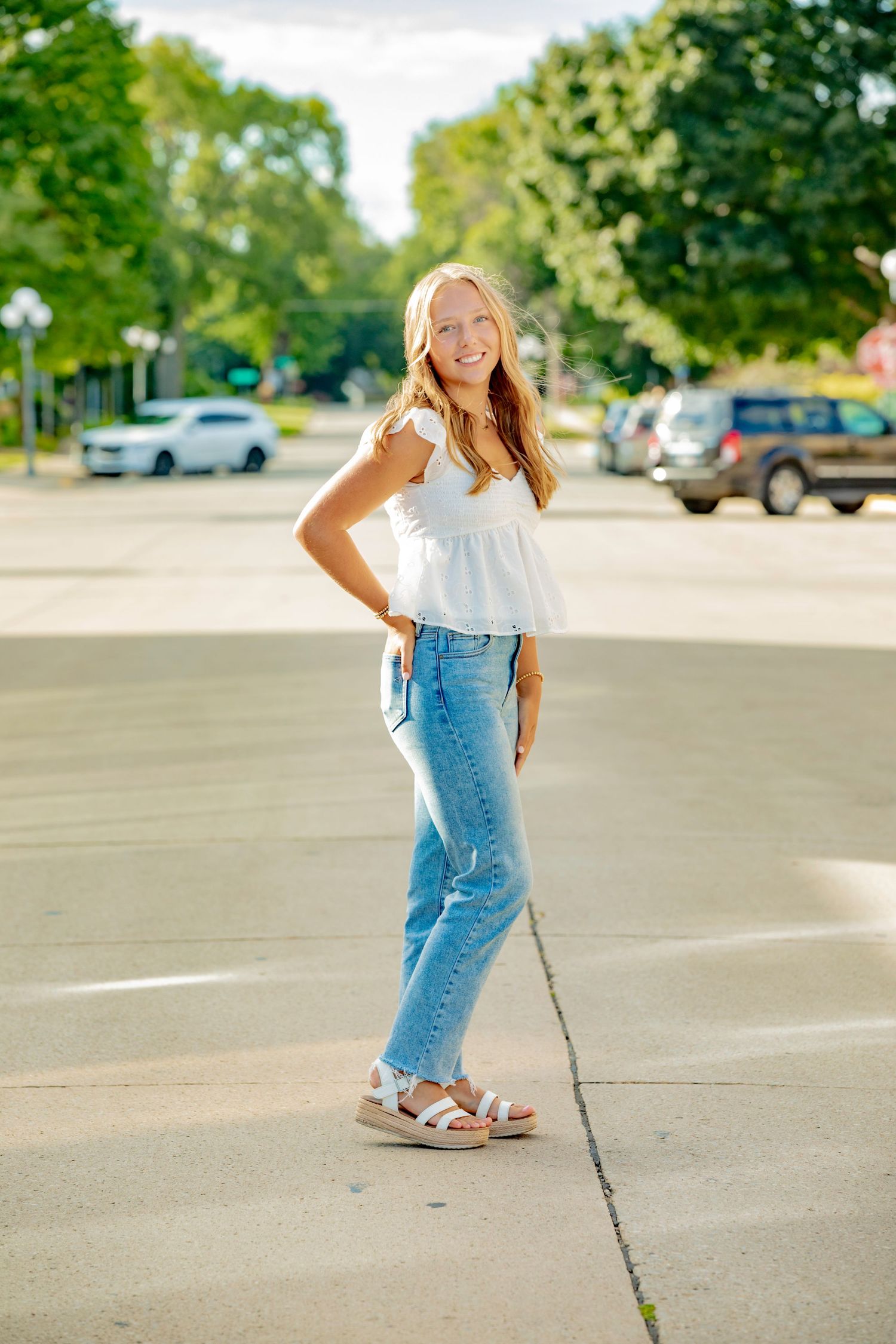 A casual streetwear look featuring light-wash denim and white sandals in an outdoor parking lot setting.