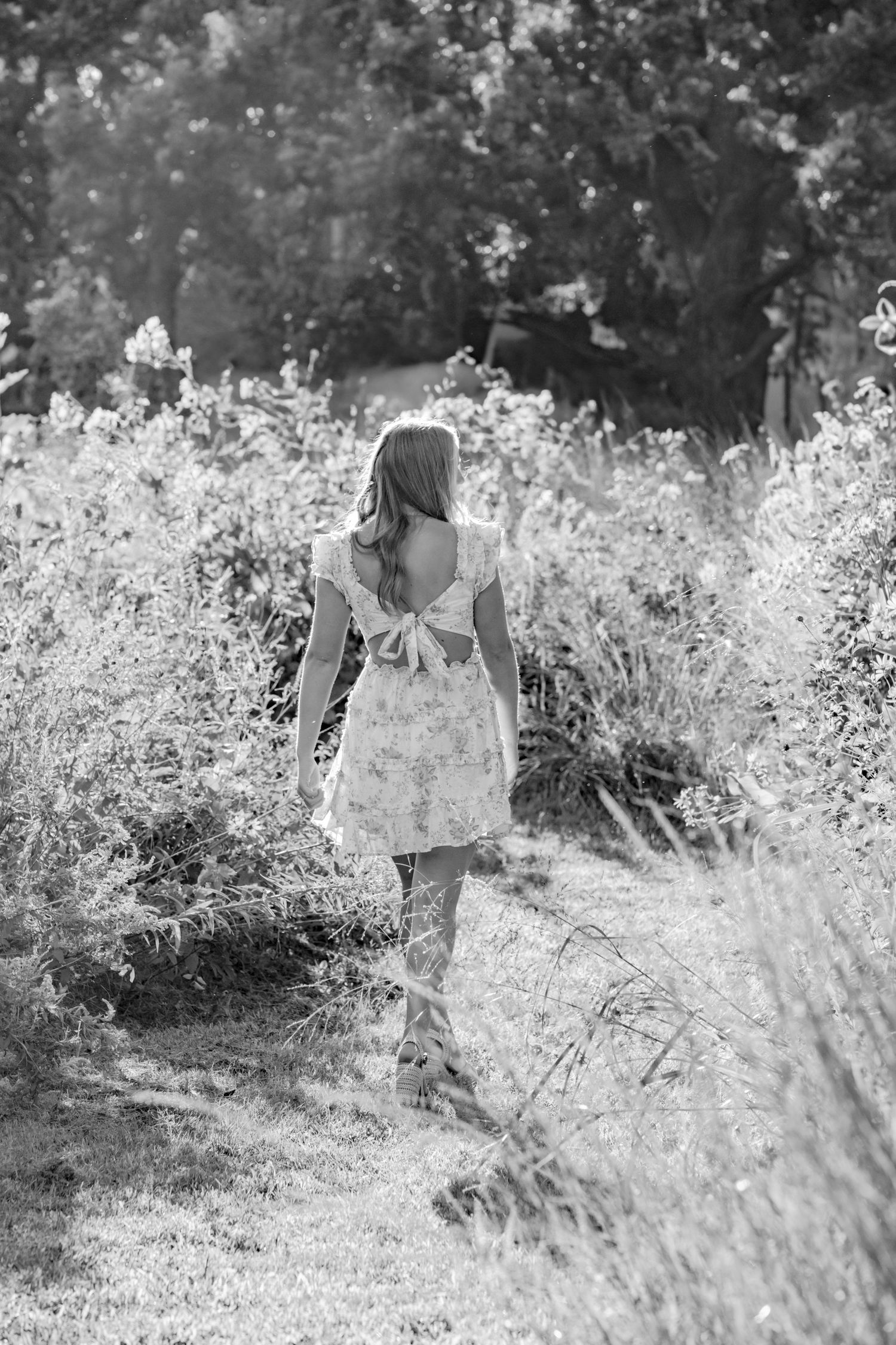 Black and white image of a figure in a light dress walking through a wildflower field on a summer day.