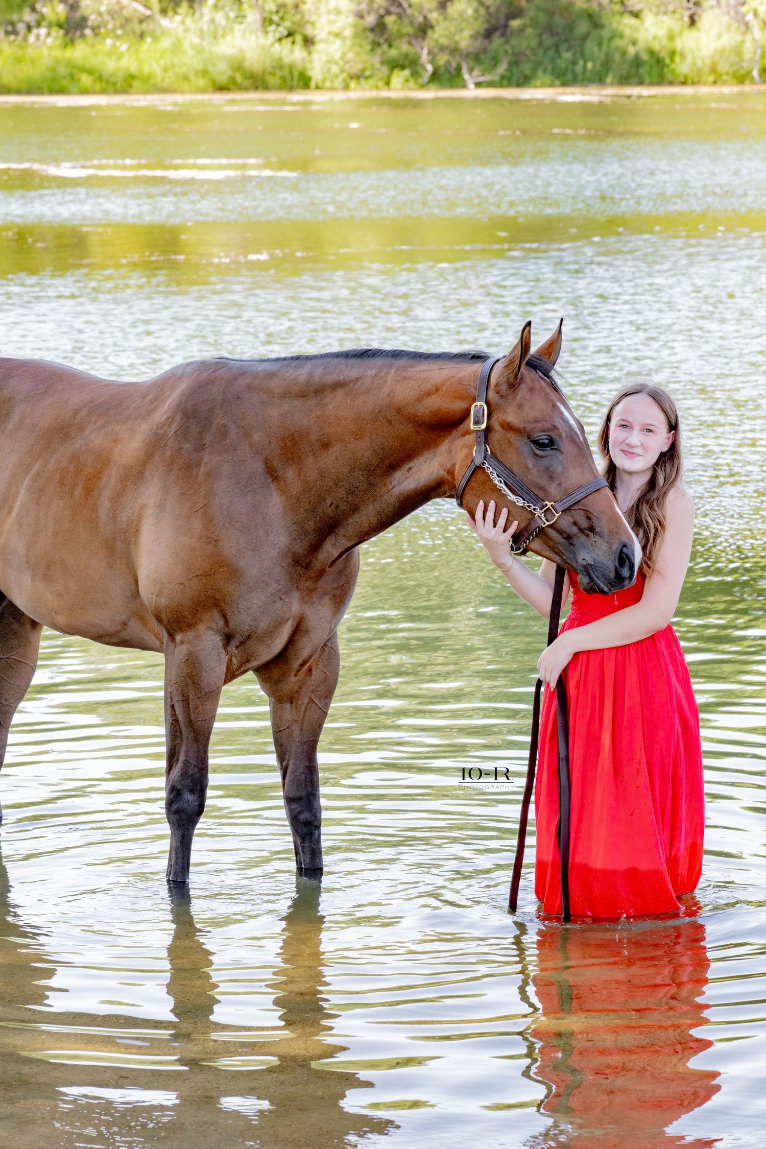 Girl in red dress standing with bay horse in shallow river water.