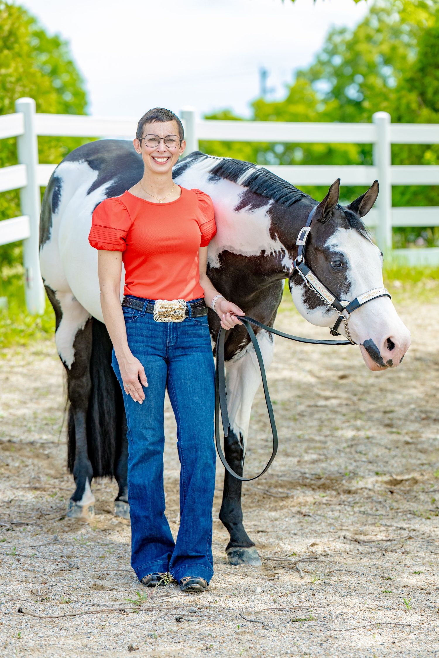 Woman in red top and jeans stands proudly beside a black and white paint horse in a sunny outdoor arena.