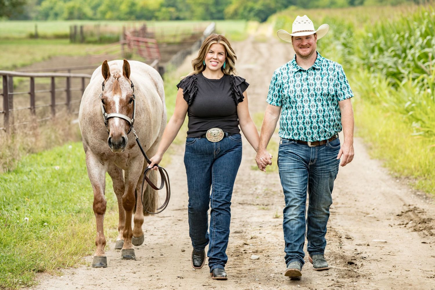 A couple walking down a dirt path with their horse on a sunny day in the countryside.