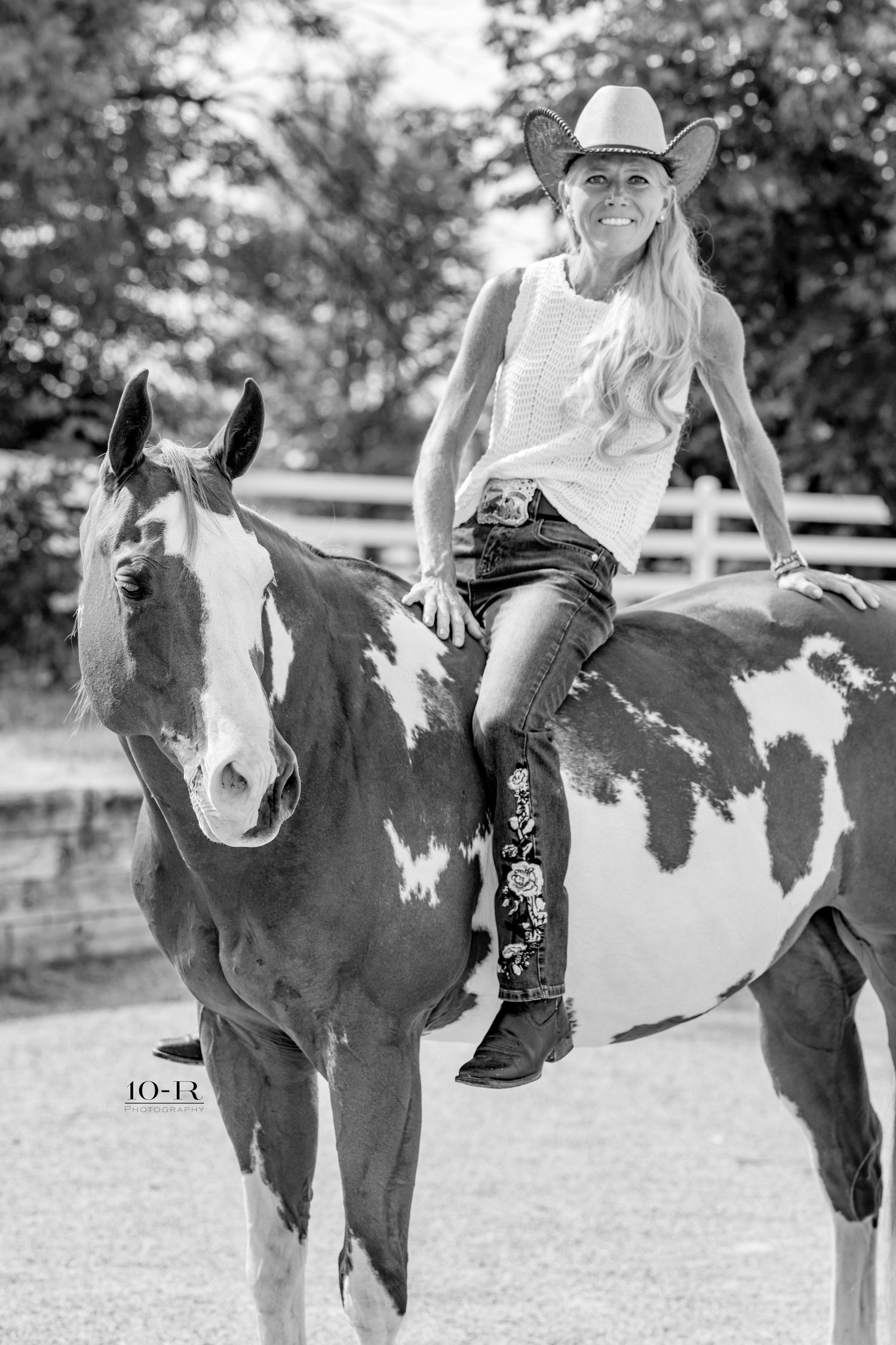 Smiling blonde woman in cowboy hat and jeans sitting atop a paint horse in a black and white outdoor photo.