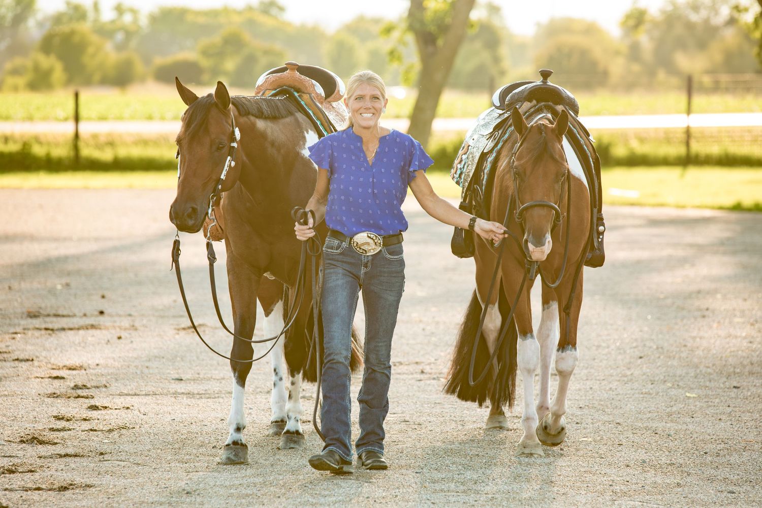 Two horses with western saddles being led down a dirt path at sunset.