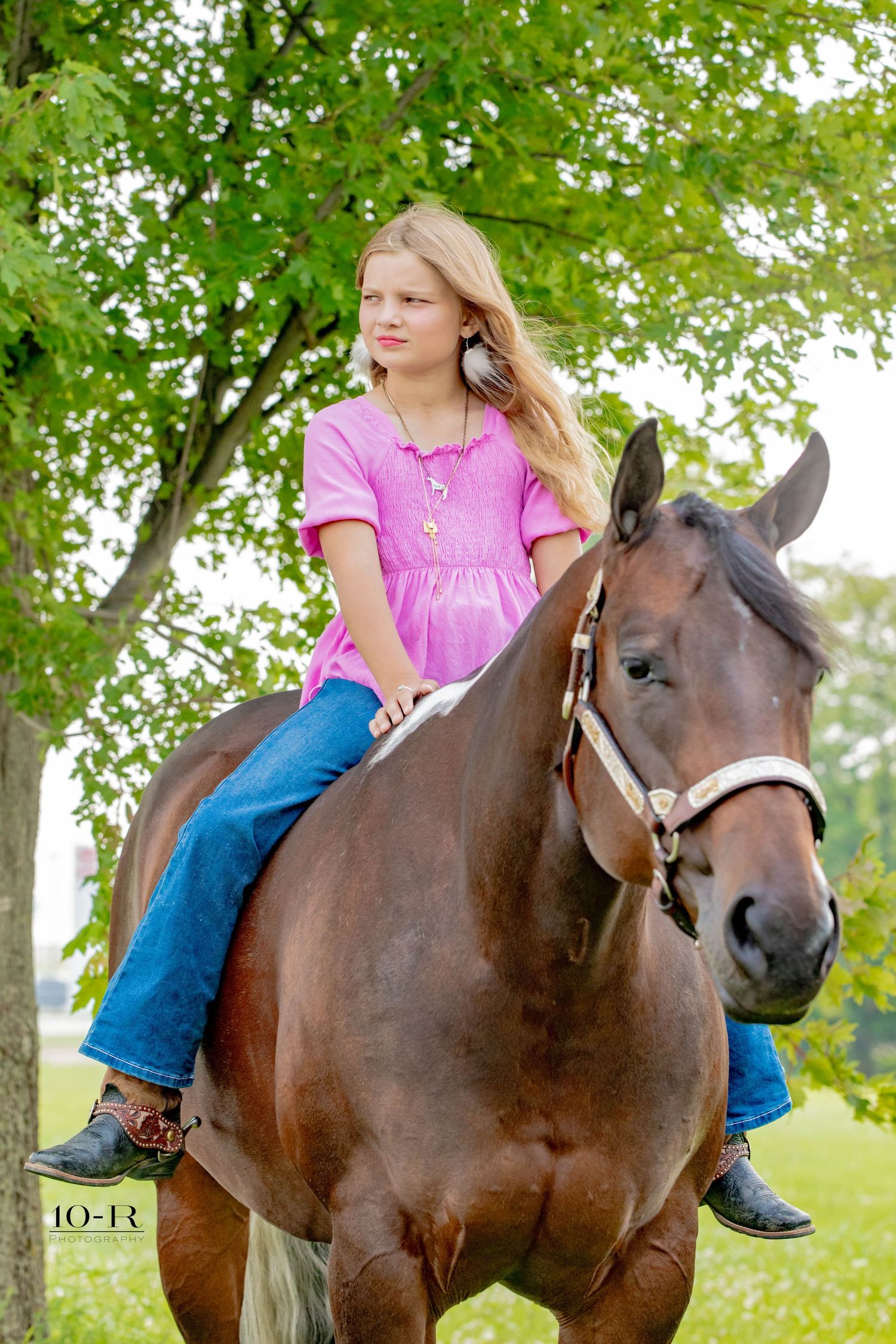 Rider in pink shirt and jeans sits atop a beautiful brown horse under green leafy trees on a sunny day.