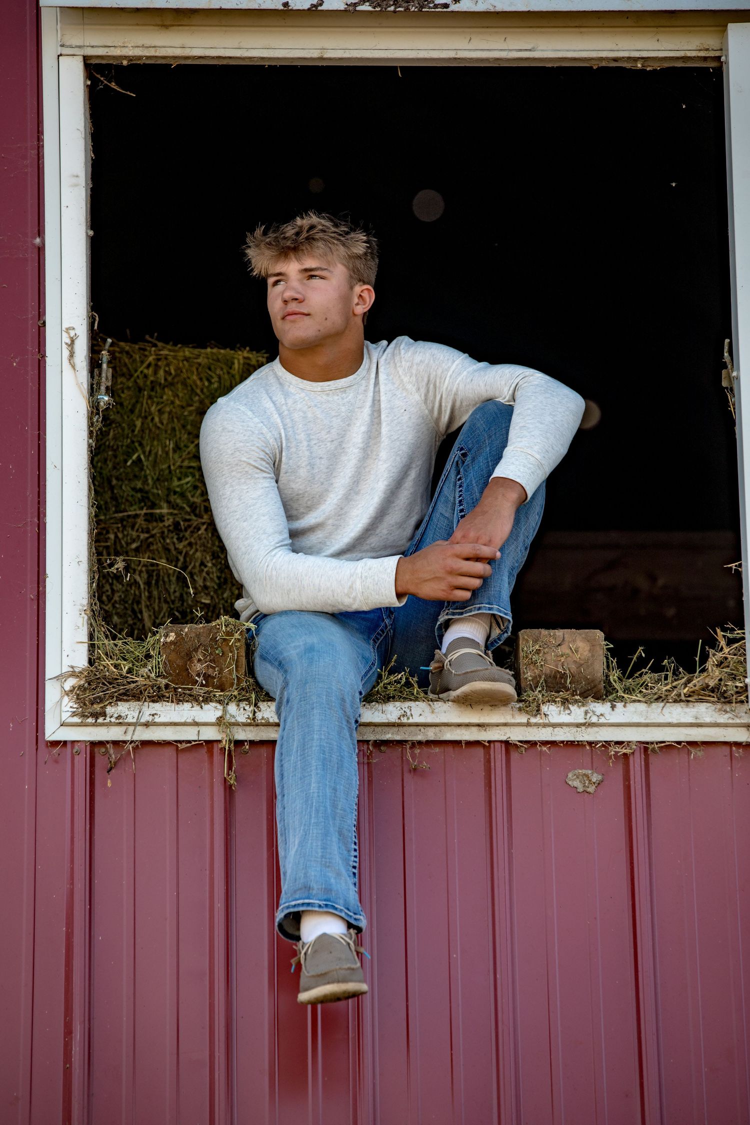 Person in light sweater and jeans sits in rustic barn window frame.