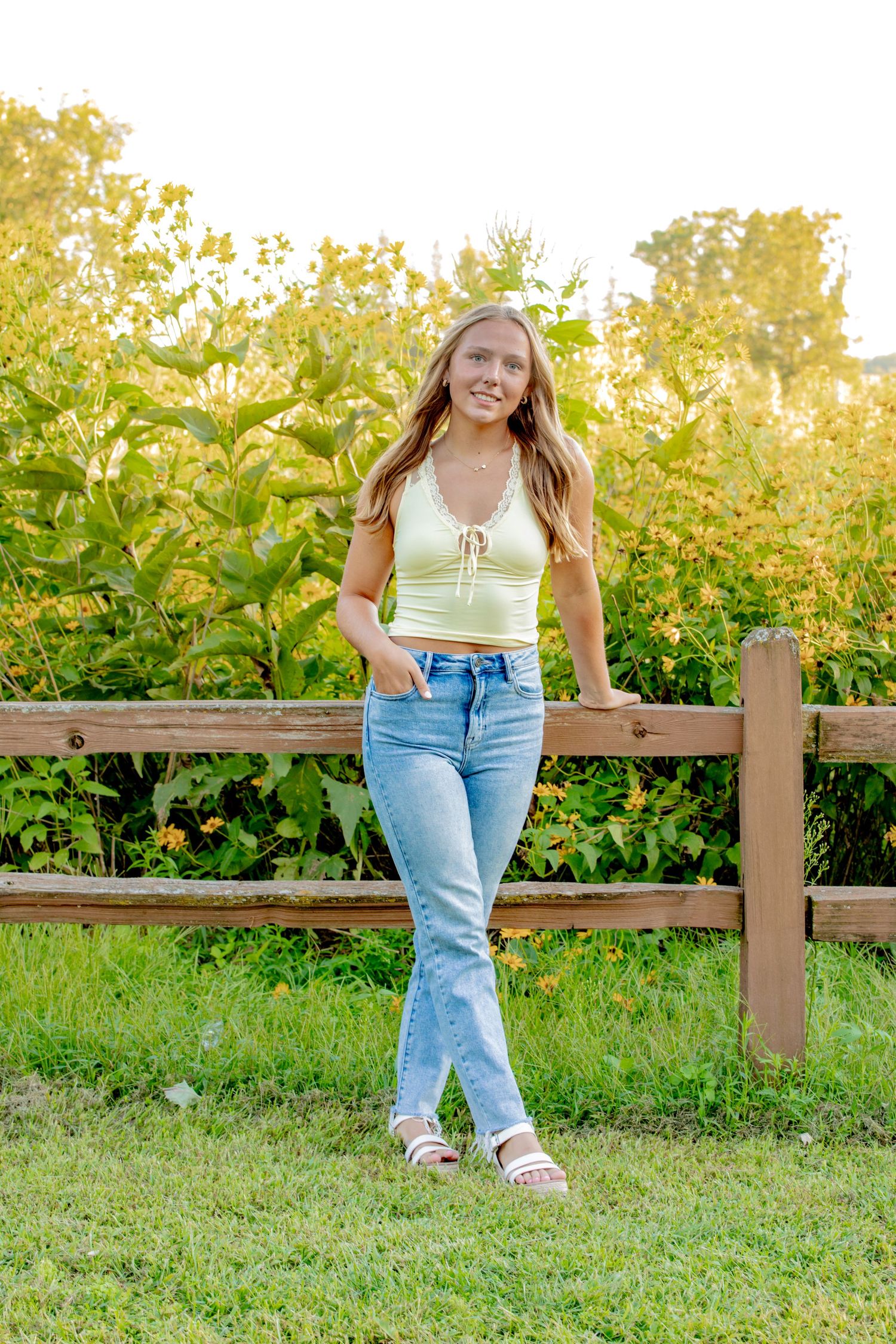 Person in casual attire sits on a wooden fence at sunset with lush greenery in the background.