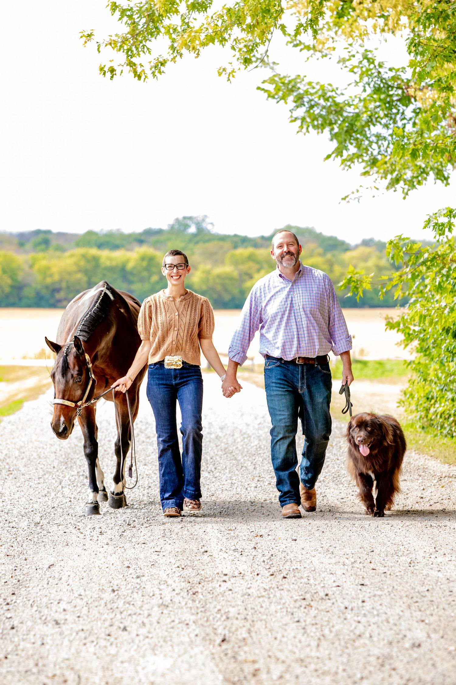 A couple walks hand in hand with their brown donkey on a gravel path surrounded by green trees and a lake view.