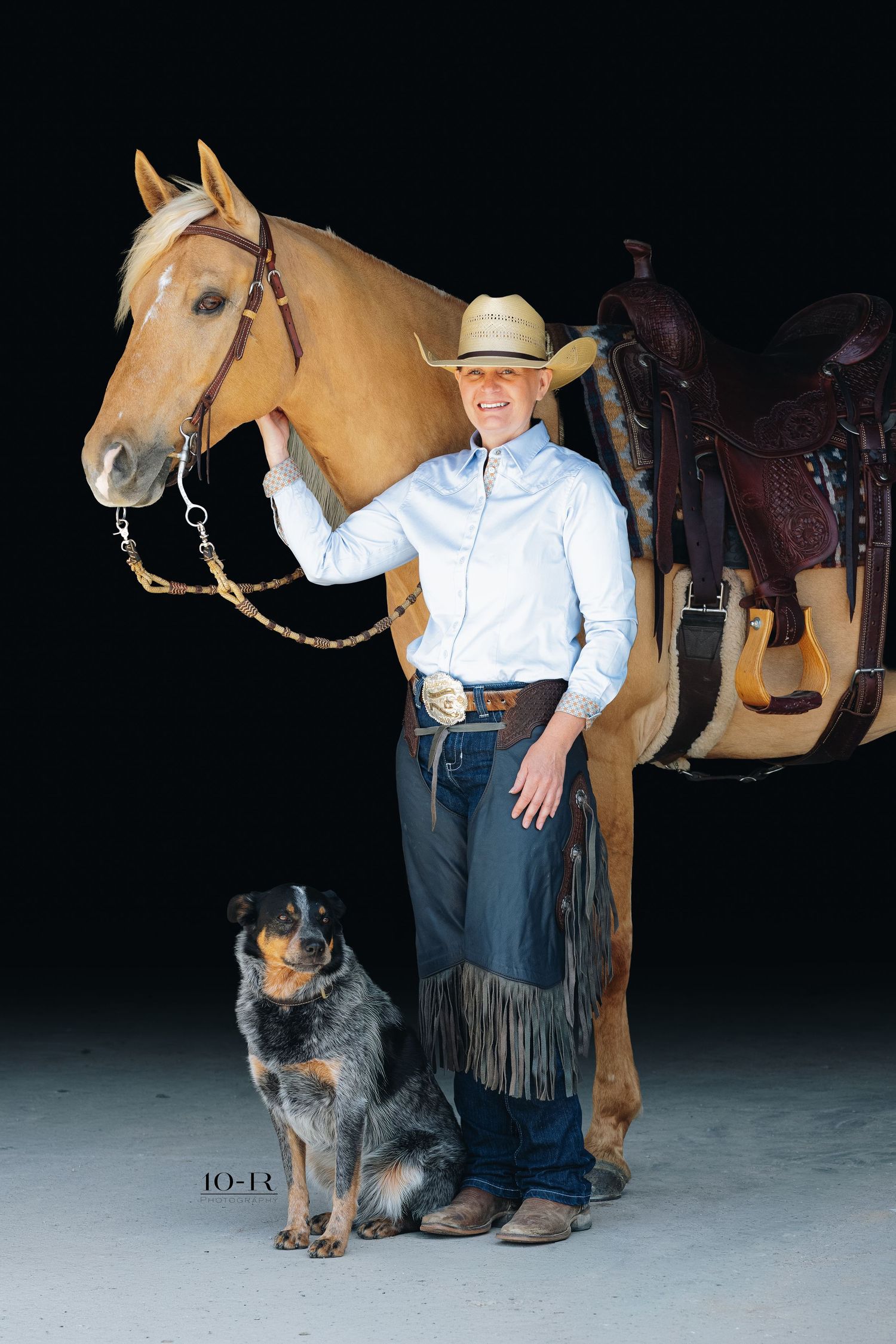 Cowboy in hat and chaps stands with a palomino horse and blue heeler dog against a dark background.